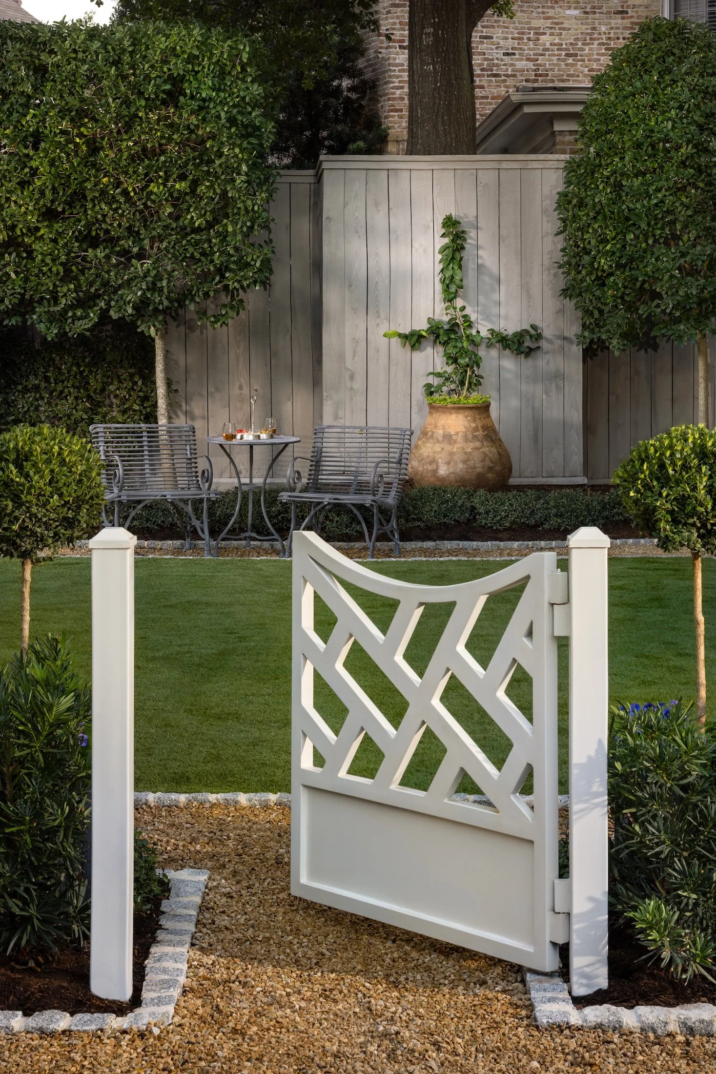 A white outdoor gate with a lattice design in a landscaped garden with green bushes, gravel path, and a large ceramic pot with a vine plant. There is a wooden fence behind the garden and a brick house wall with a tree growing nearby.