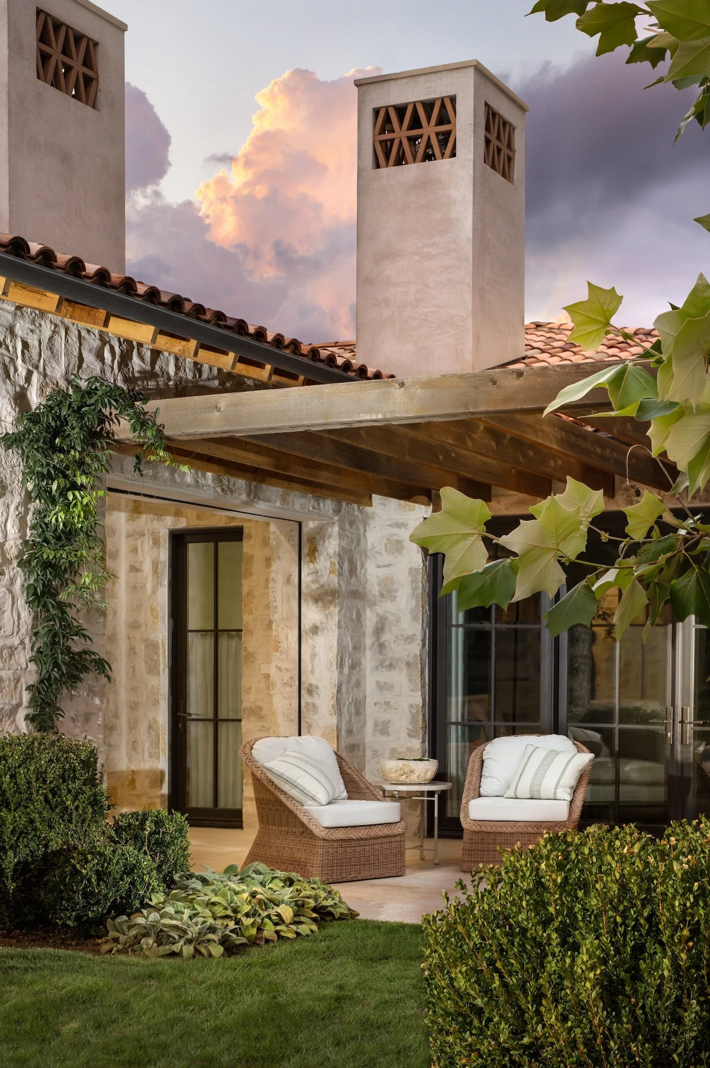 A cozy outdoor patio area with two wicker chairs and cushions next to a large glass door, surrounded by lush green bushes and plants, with a stone building wall and a chimney in the background, under a partly cloudy sky at sunset.