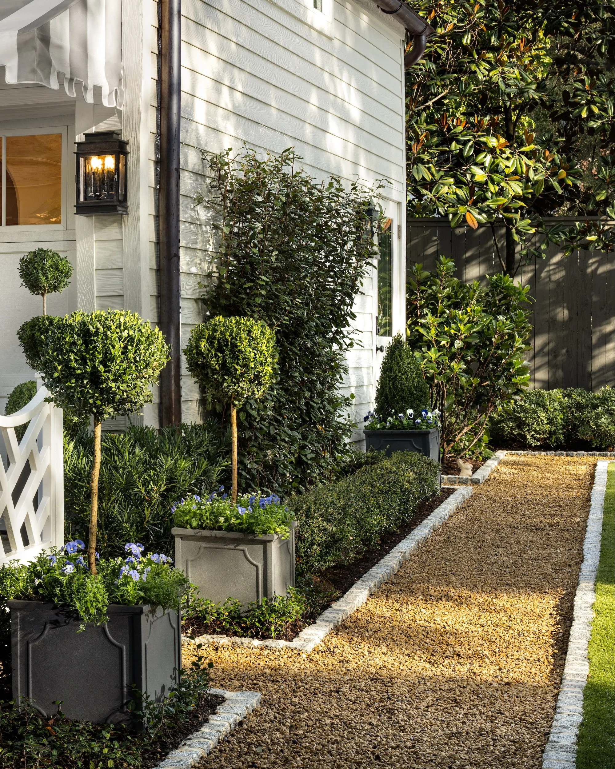 A garden pathway with small green trees and flowering plants in decorative pots next to a white house wall, with sunlight casting shadows and a gravel walkway bordered by stones.