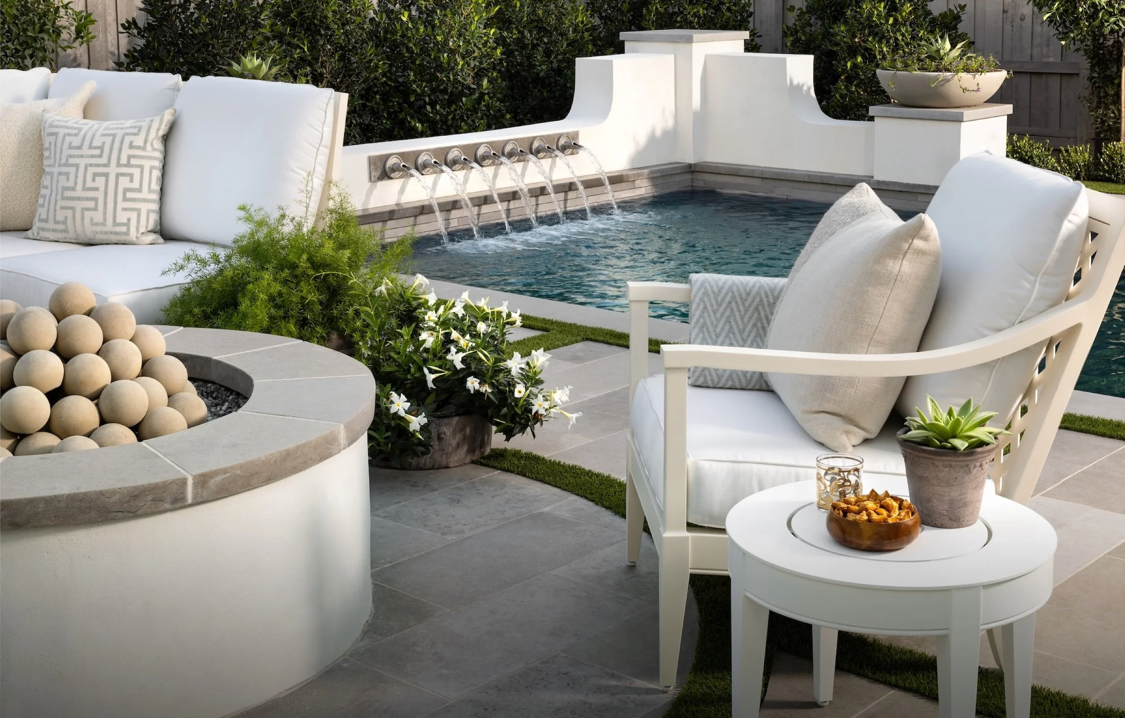 A backyard patio with white cushioned patio furniture, potted plants, a fire pit filled with beige ceramic balls, and a small pool with water flowing from spouts on a white wall, with greenery and a wooden fence in the background.