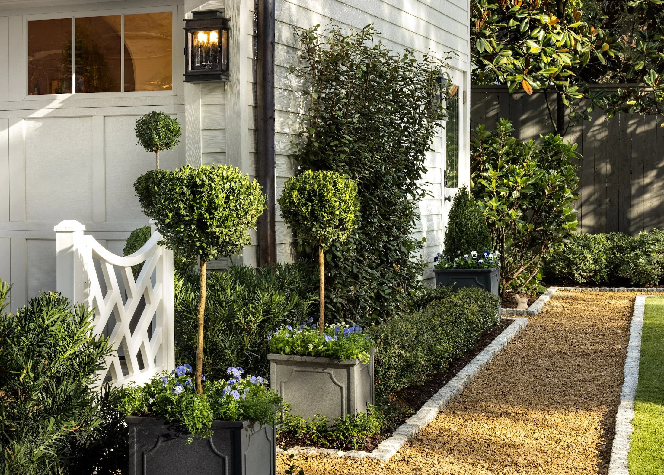 Front yard garden with trimmed bushes, potted plants, a white lattice fence, a house wall with a lantern, and a gravel pathway