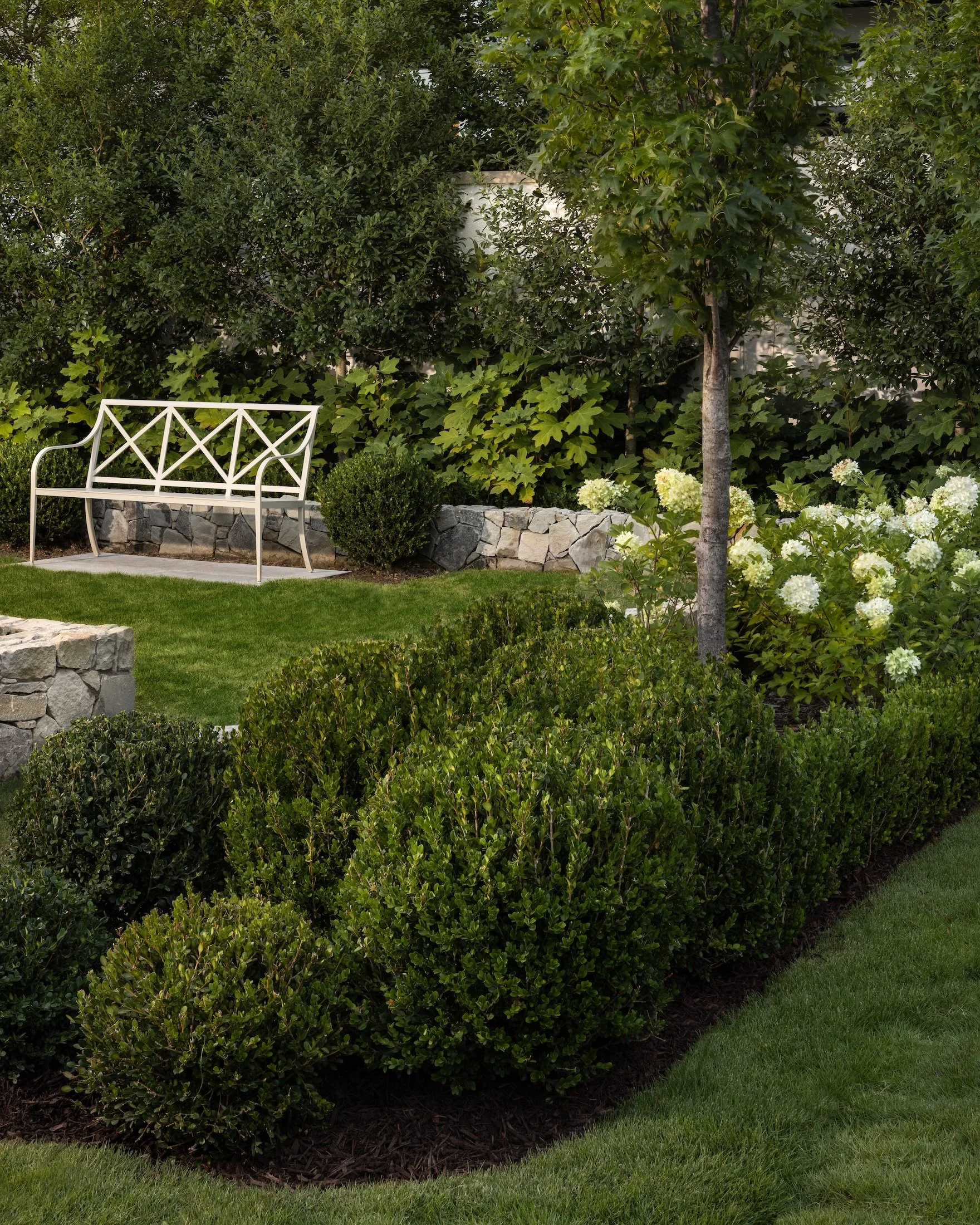 A garden scene with a white metal park bench, lush green bushes, one small tree, and a flower bed with white hydrangeas on the side, bordered by a stone wall and grass.