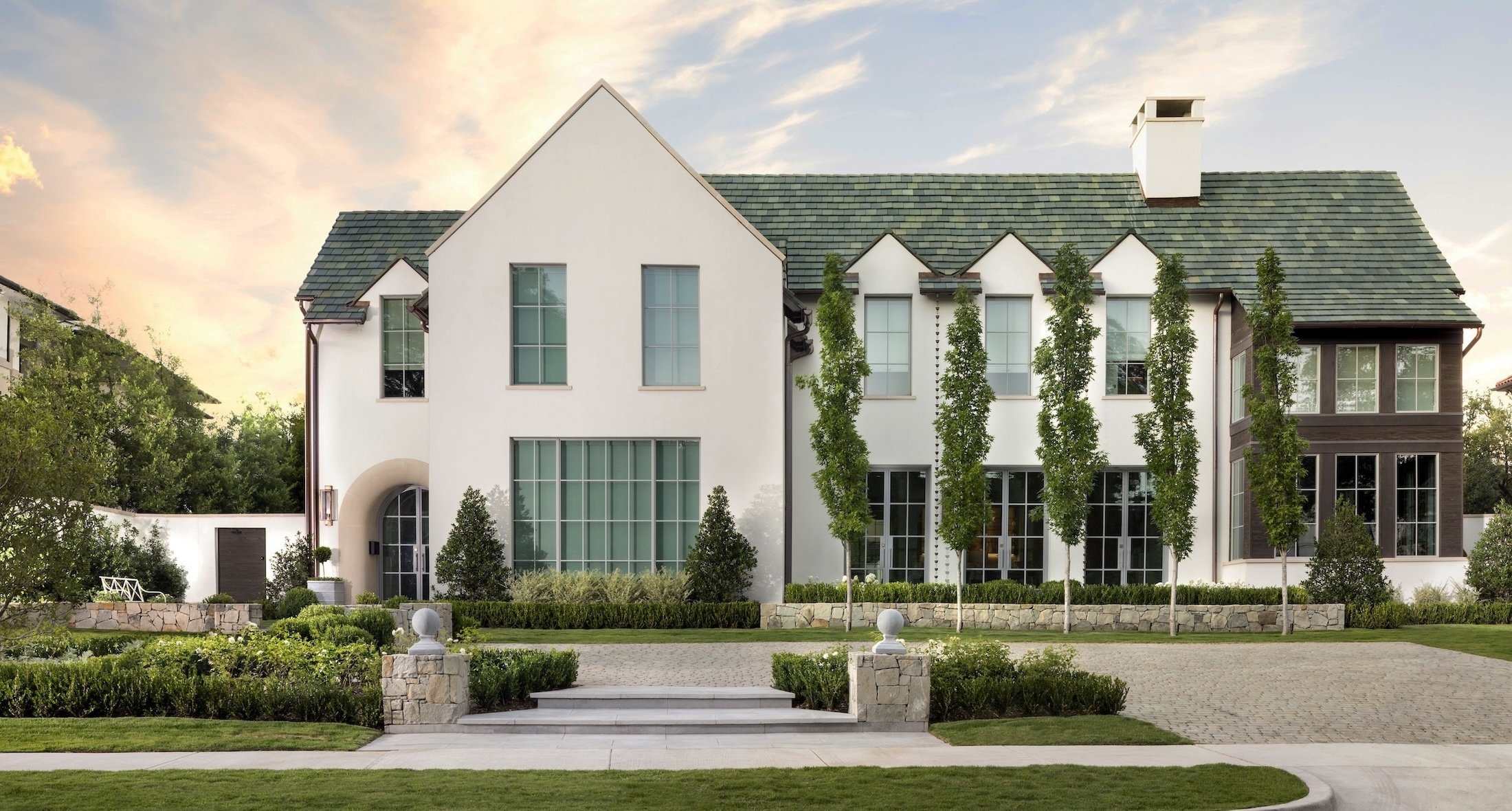 A modern white house with multiple large windows and a green tiled roof, surrounded by a well-manicured lawn, small trees, and landscaping elements underneath a partly cloudy sky.