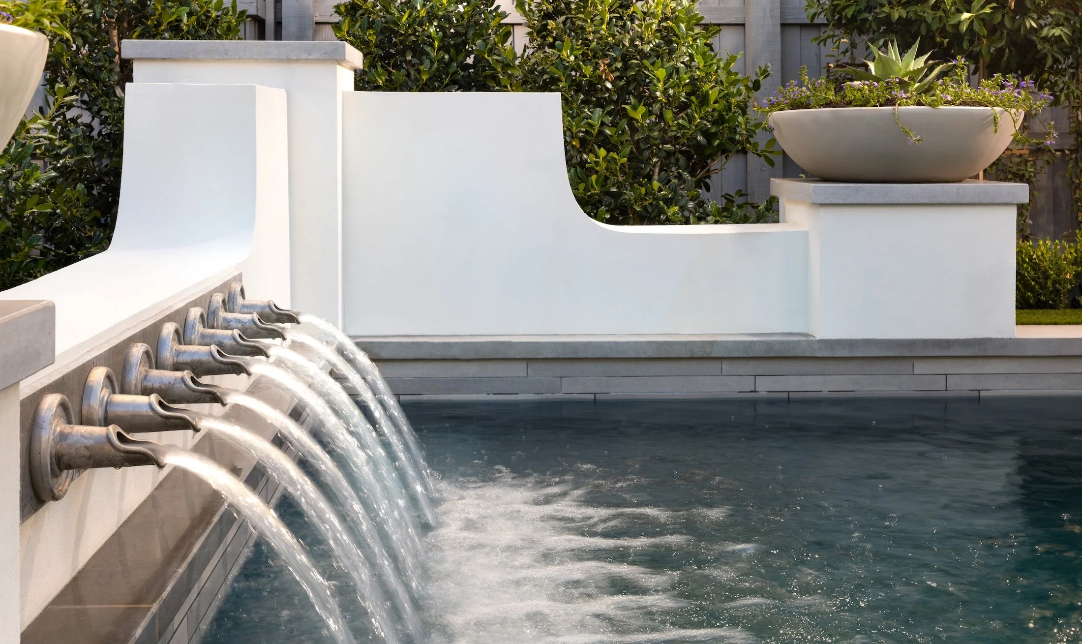 Close-up of a modern water fountain with multiple streams of water flowing from metal spouts into a pool, with a white wall and greenery in the background.