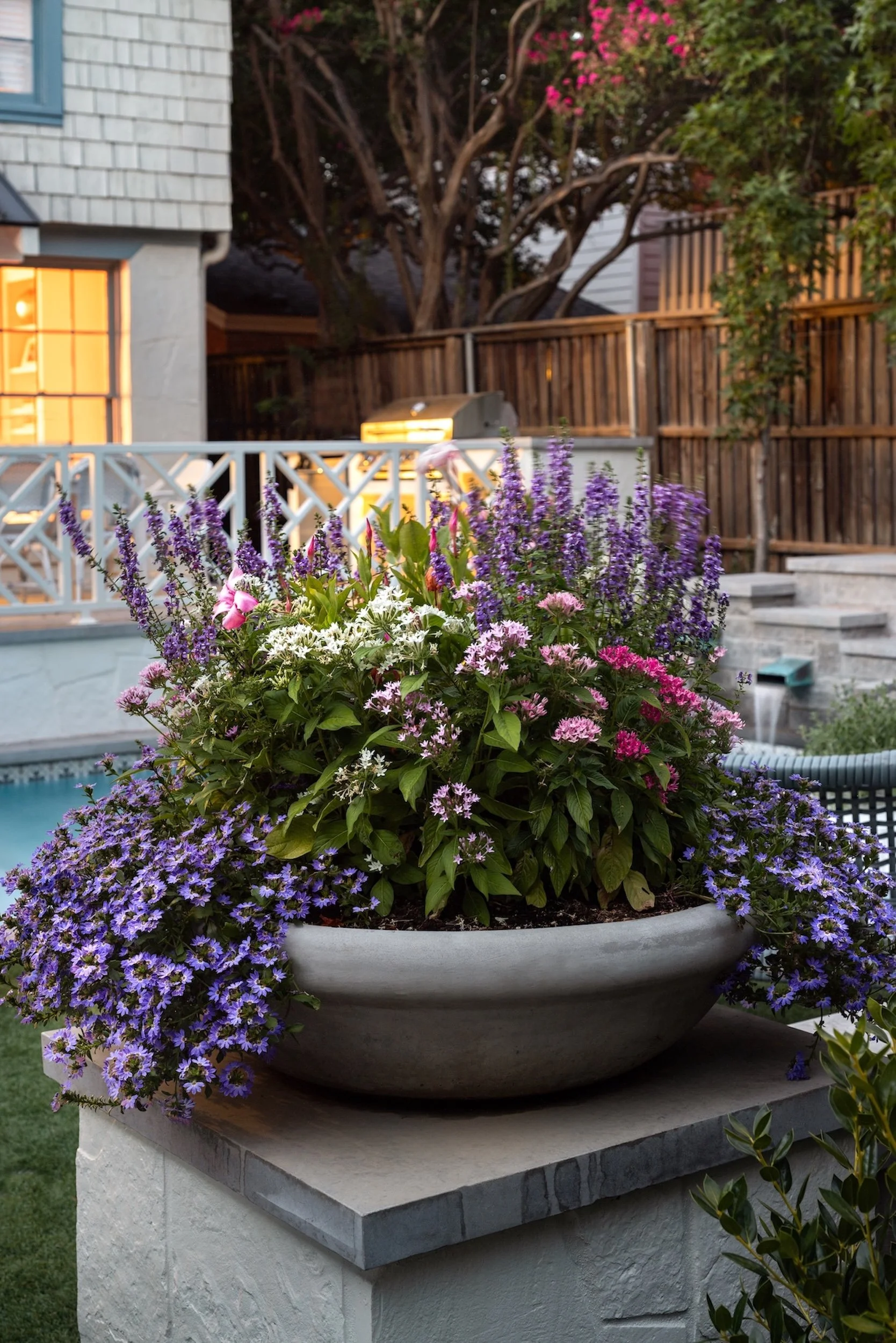 A large white planter filled with purple, pink, and white flowers sits on a stone ledge in a backyard. In the background, there is a wooden fence, a grill, and a large tree with pink flowers.