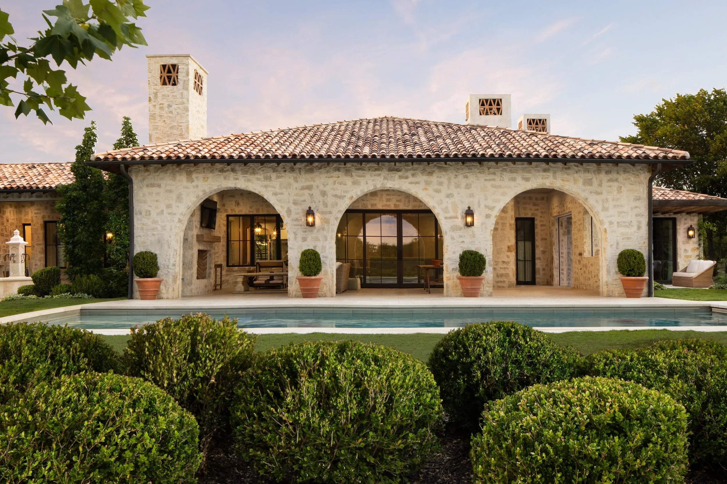 A house made of beige stone with a terracotta tile roof, arched porch, and large windows reflecting the sky. There are three chimney stacks with decorative vents. The house is surrounded by manicured bushes and a pool in the foreground, with trees and a partly cloudy sky in the background.
