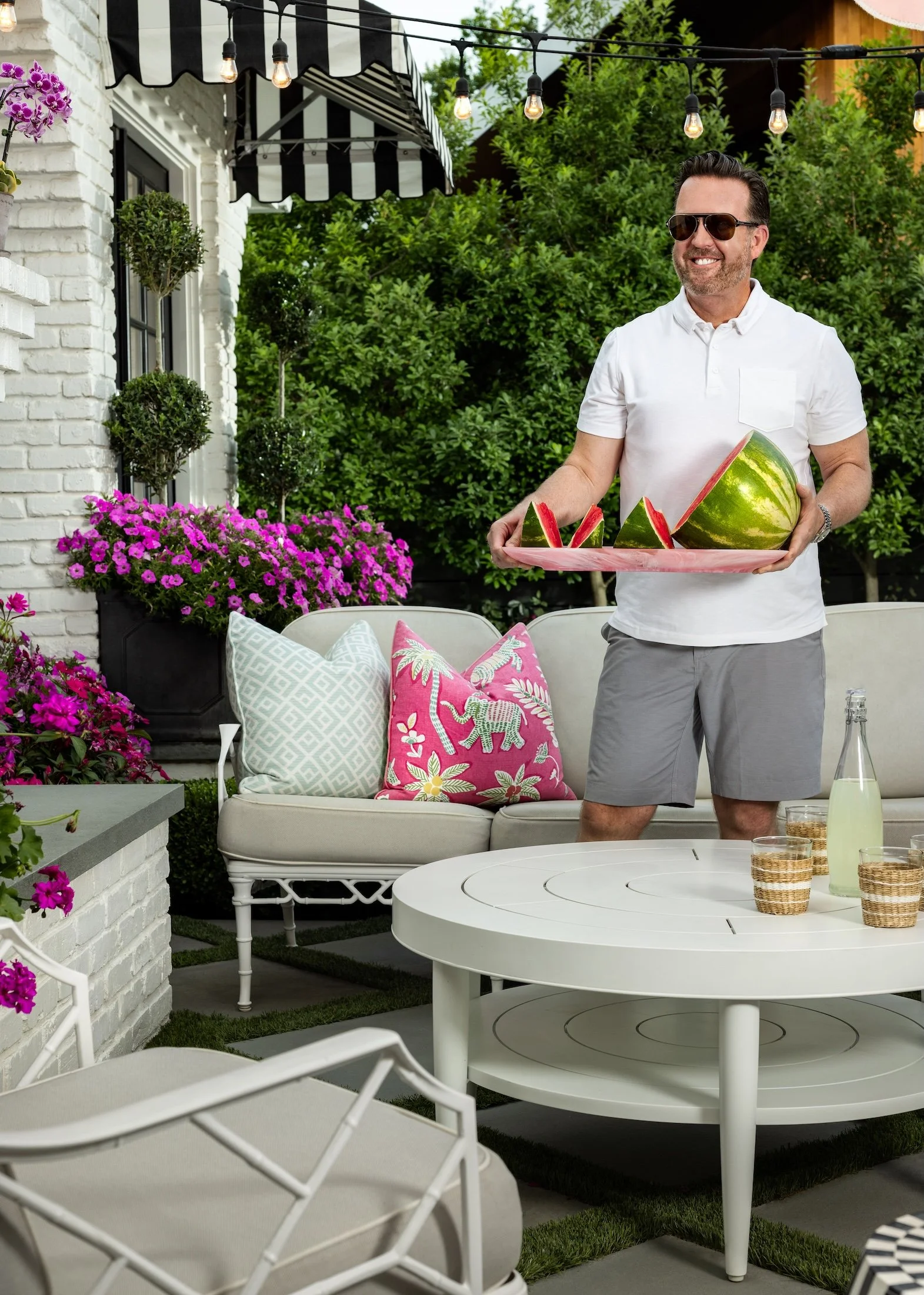 A man in sunglasses and a white polo shirt holds a tray with slices of watermelon in an outdoor patio with white furniture, pink and white pillows, potted flowers, and string lights.
