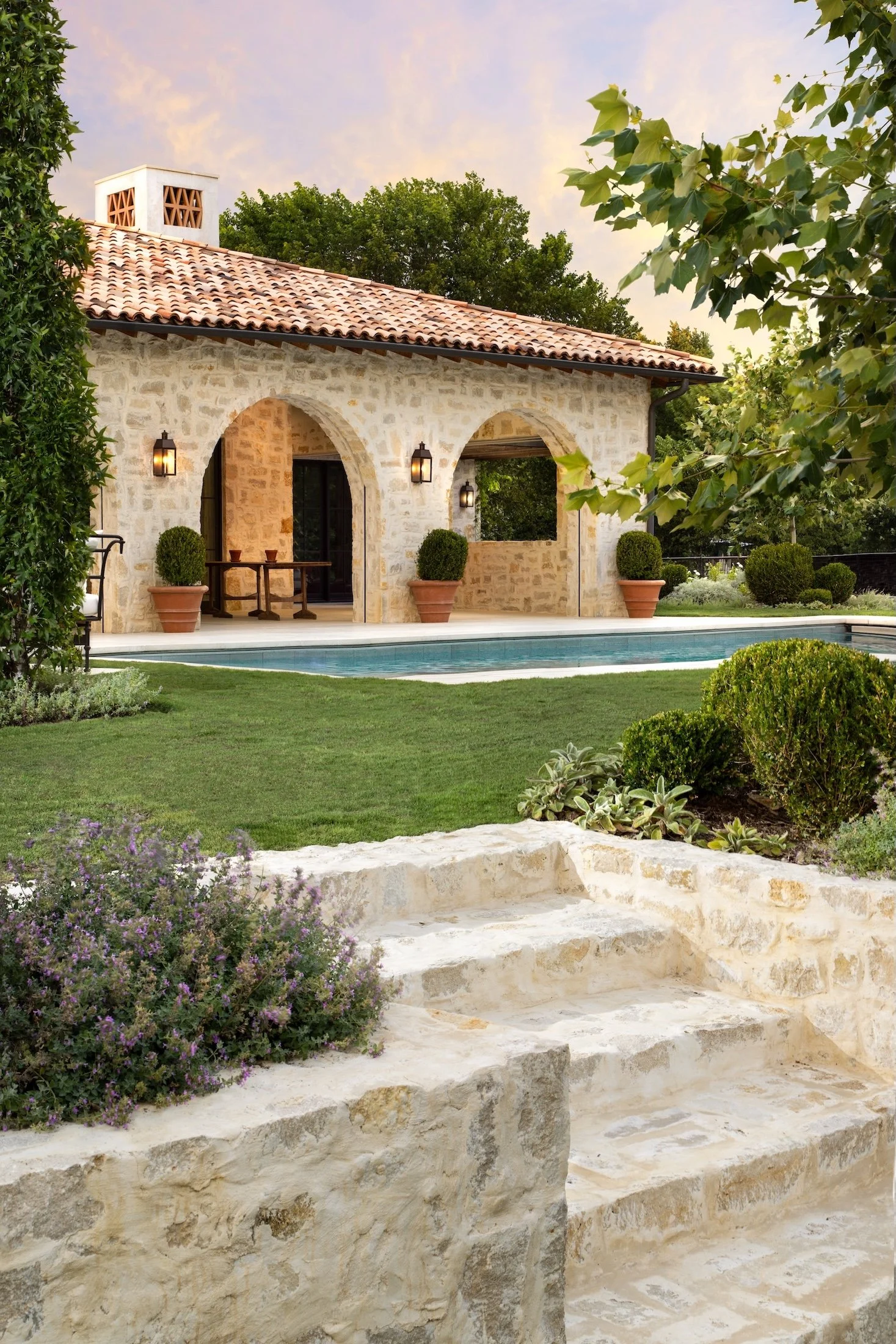 A stone house with a red tile roof, arched doorways, outdoor wall lanterns, and potted plants in a lush garden with a swimming pool in the foreground, under a pastel sky.
