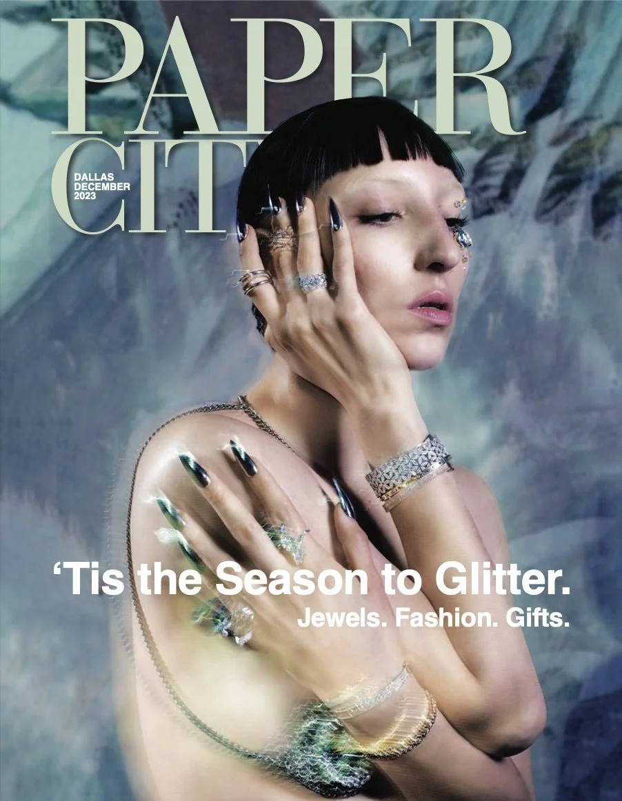 A woman with short black hair and multiple rings, bracelets, and necklaces, touching her face and shoulder, against a background of abstract art, with the text 'PAPER CITY DALLAS DECEMBER 2023' and a caption that reads 'Tis the Season to Glitter. Jewels. Fashion. Gifts.'.