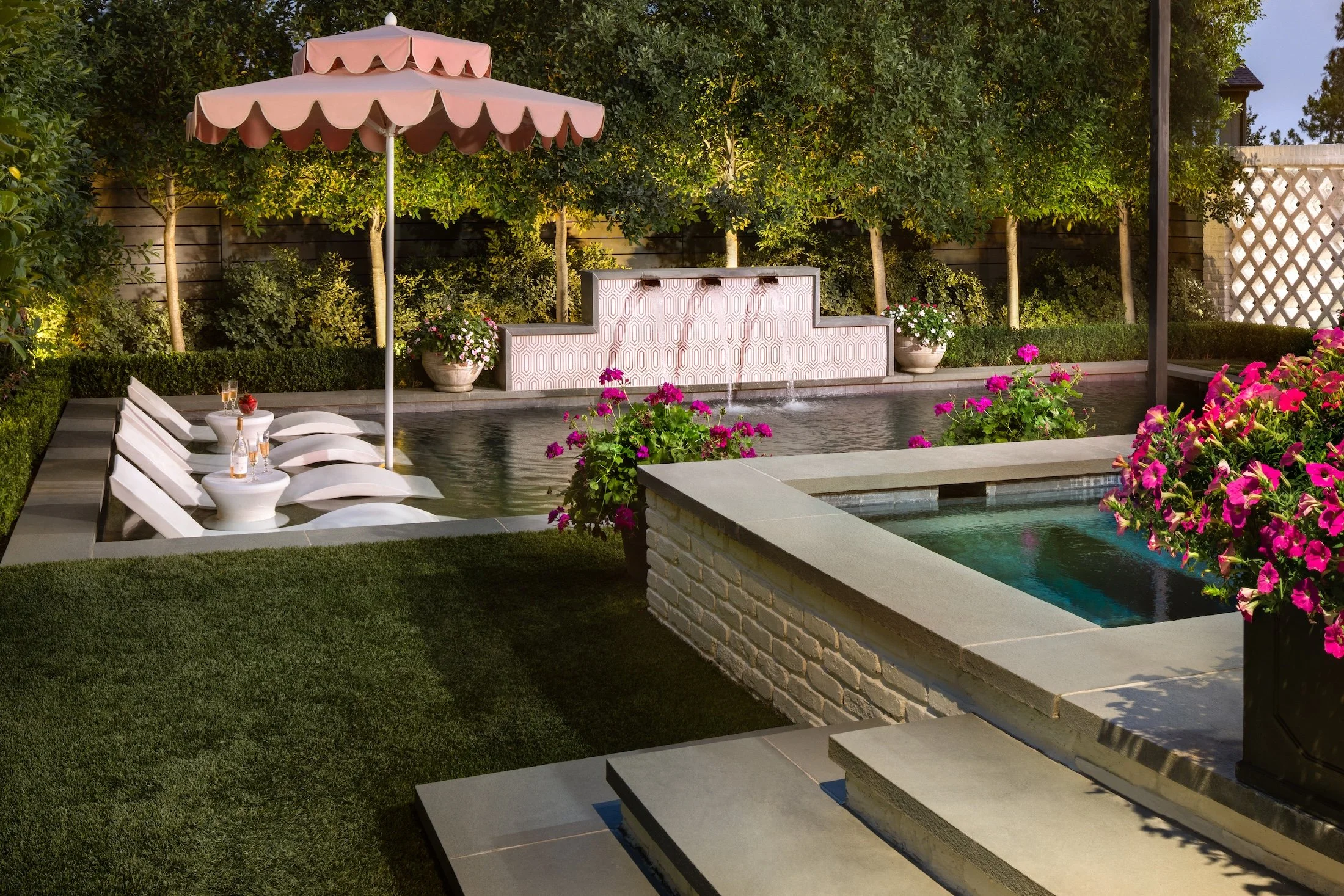 A backyard pool area with lounge chairs, a pink umbrella, and lush green trees and plants. There are decorative planters with flowers and a small fountain feature in the pool.