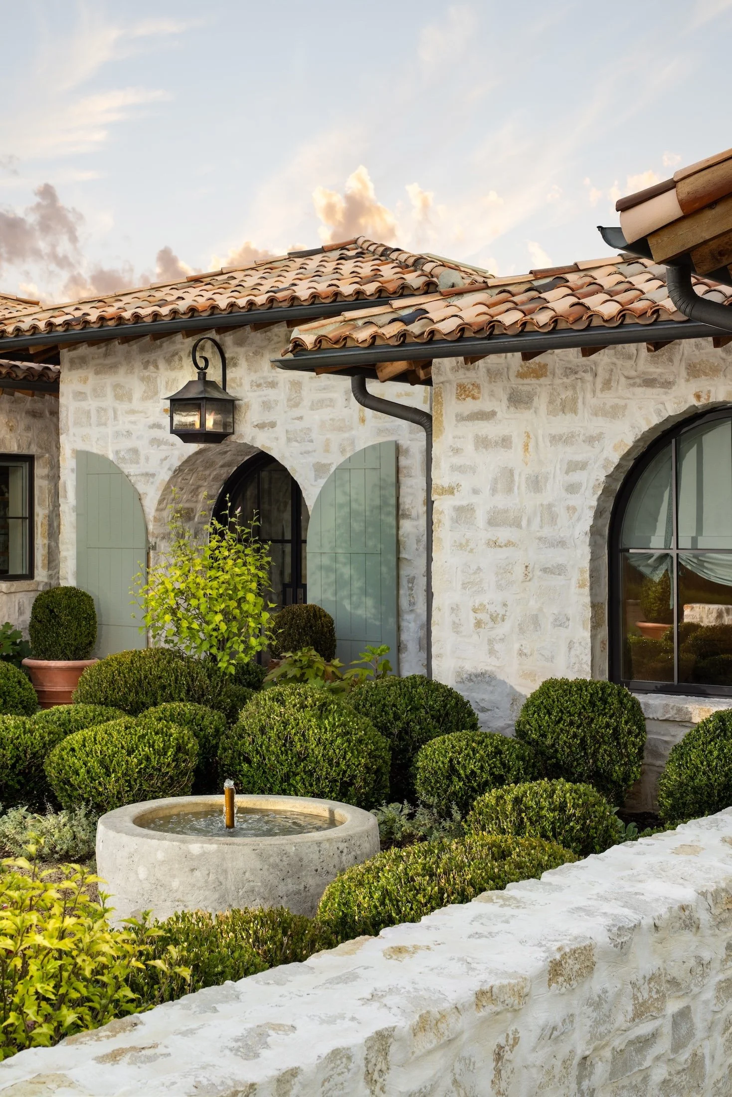 A stone house with a tiled roof, arched windows, and a black lantern outside. The garden features neatly trimmed bushes, a small fountain, and potted plants, with a cloudy sky in the background.