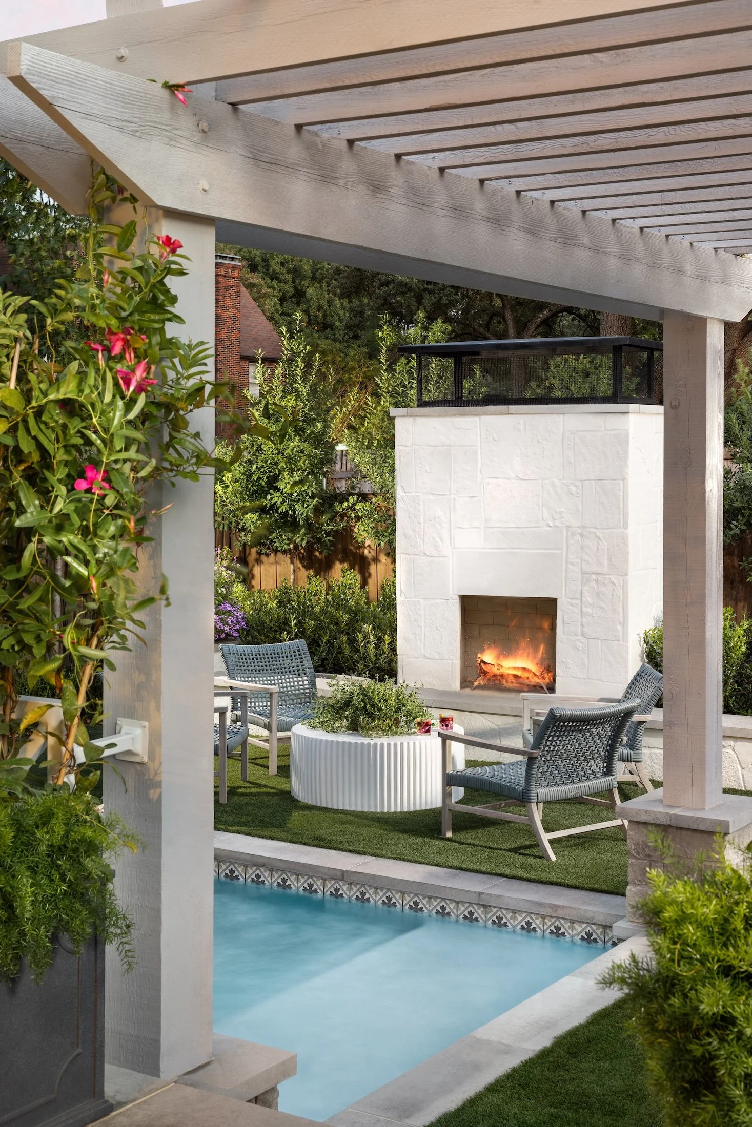 A backyard patio with a rectangular fire pit, surrounded by four gray outdoor chairs, adjacent to a swimming pool with decorative tiles at the edge. There's a pergola overhead, and lush greenery and shrubs around the area, with a house and trees in the background.