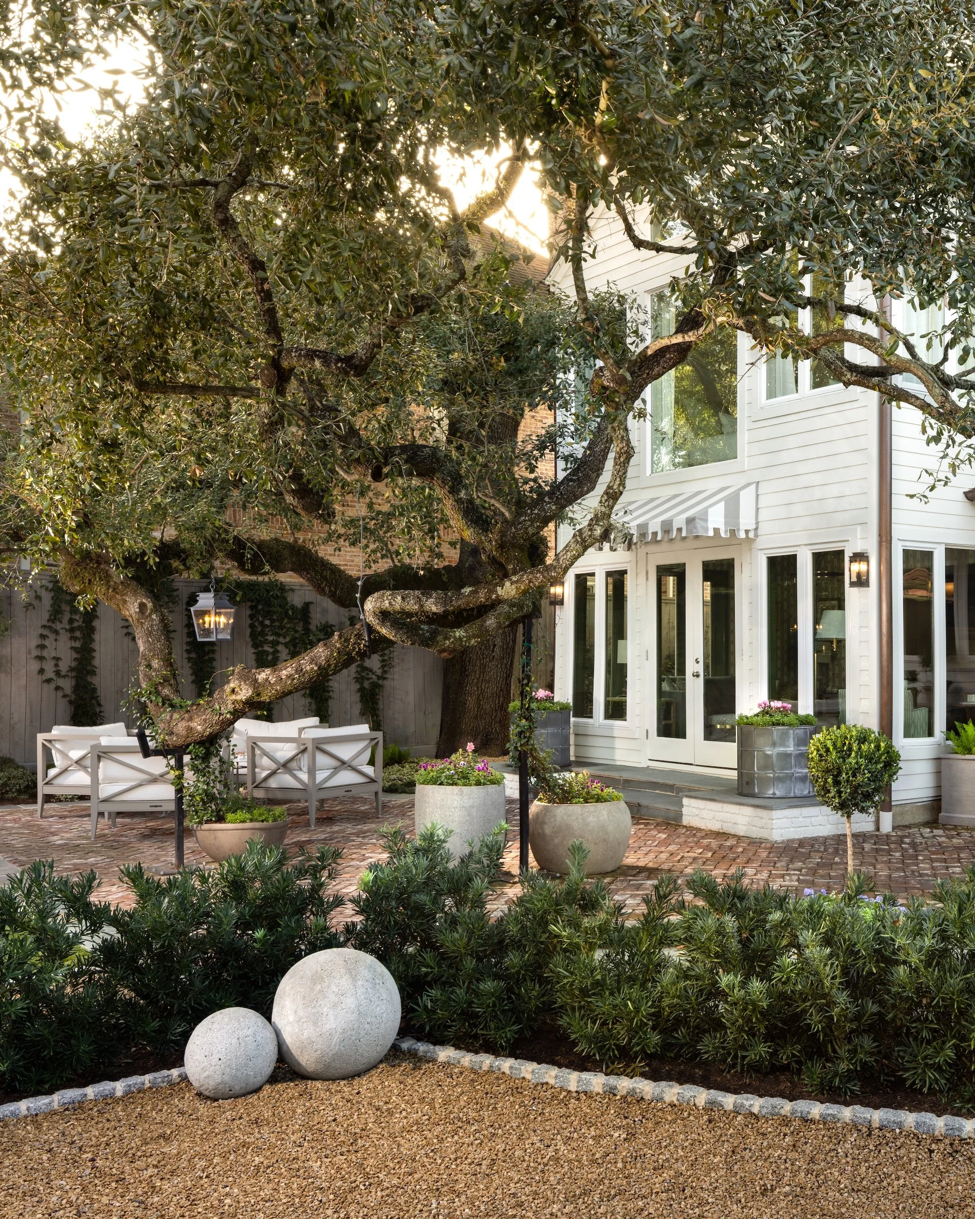 A well-maintained backyard with a large tree, outdoor seating area, and potted plants in front of a white house with large windows.