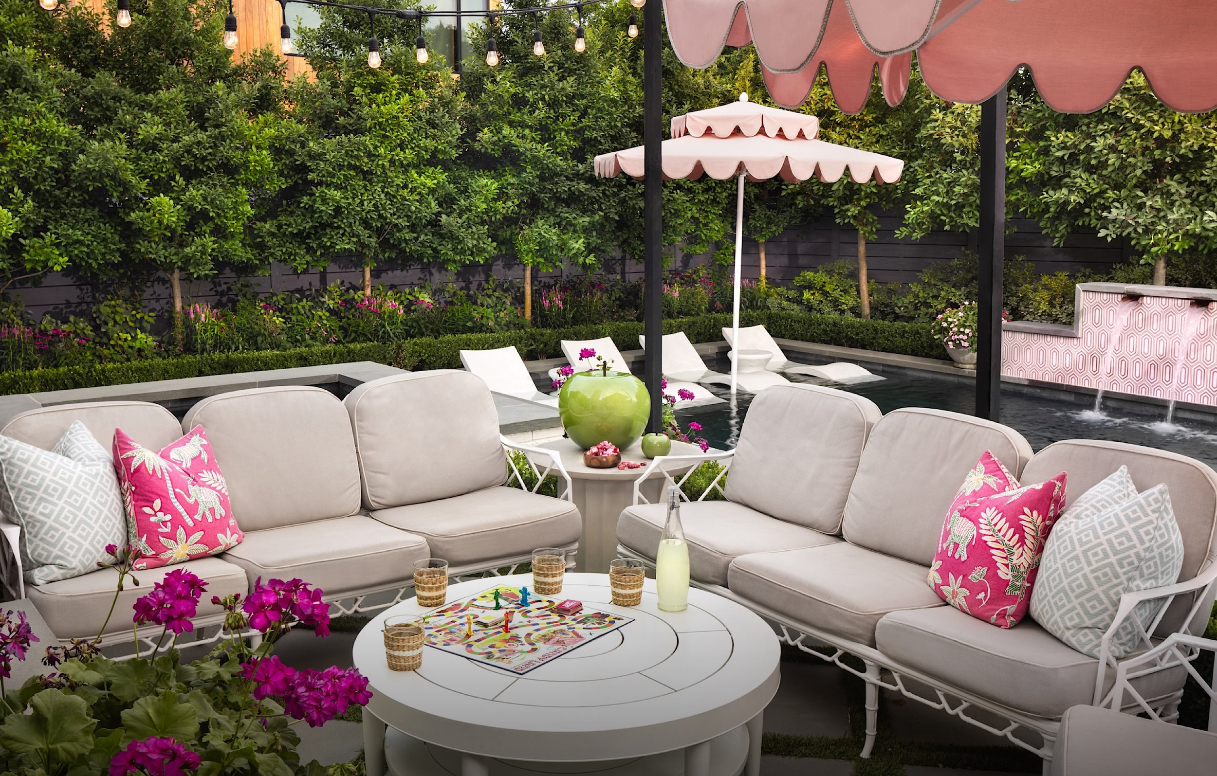 Outdoor patio with white sofa and pink, white, and patterned pillows, a circular white table with colorful board game, drinks, and small glasses, surrounded by pink flowers. In the background, there are green bushes, a small pool with pink umbrellas, lounge chairs, and string lights hanging overhead.
