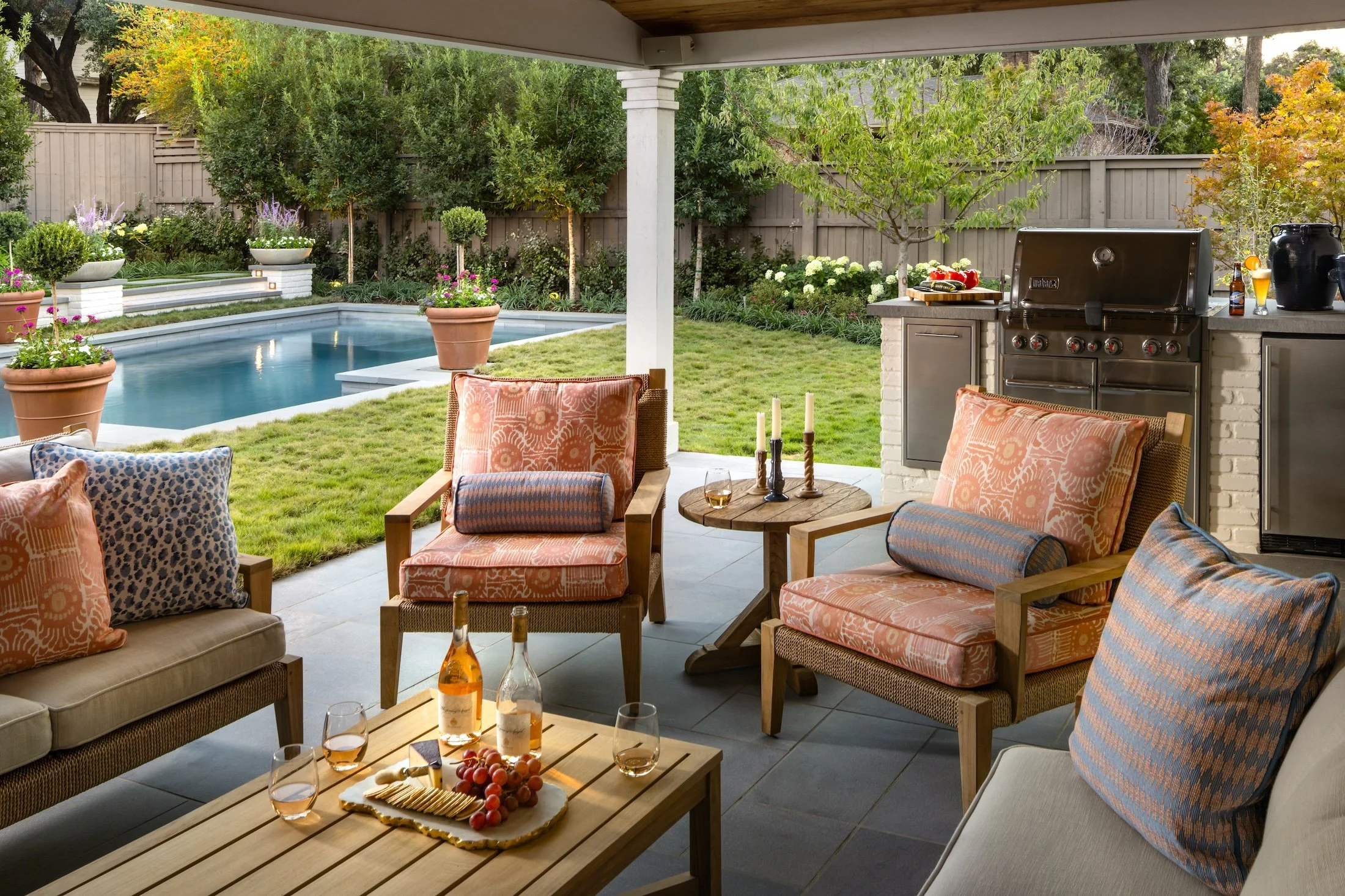Patio with outdoor furniture, a swimming pool, and garden with trees and flowering plants, outdoor grill, and drinks on a table.
