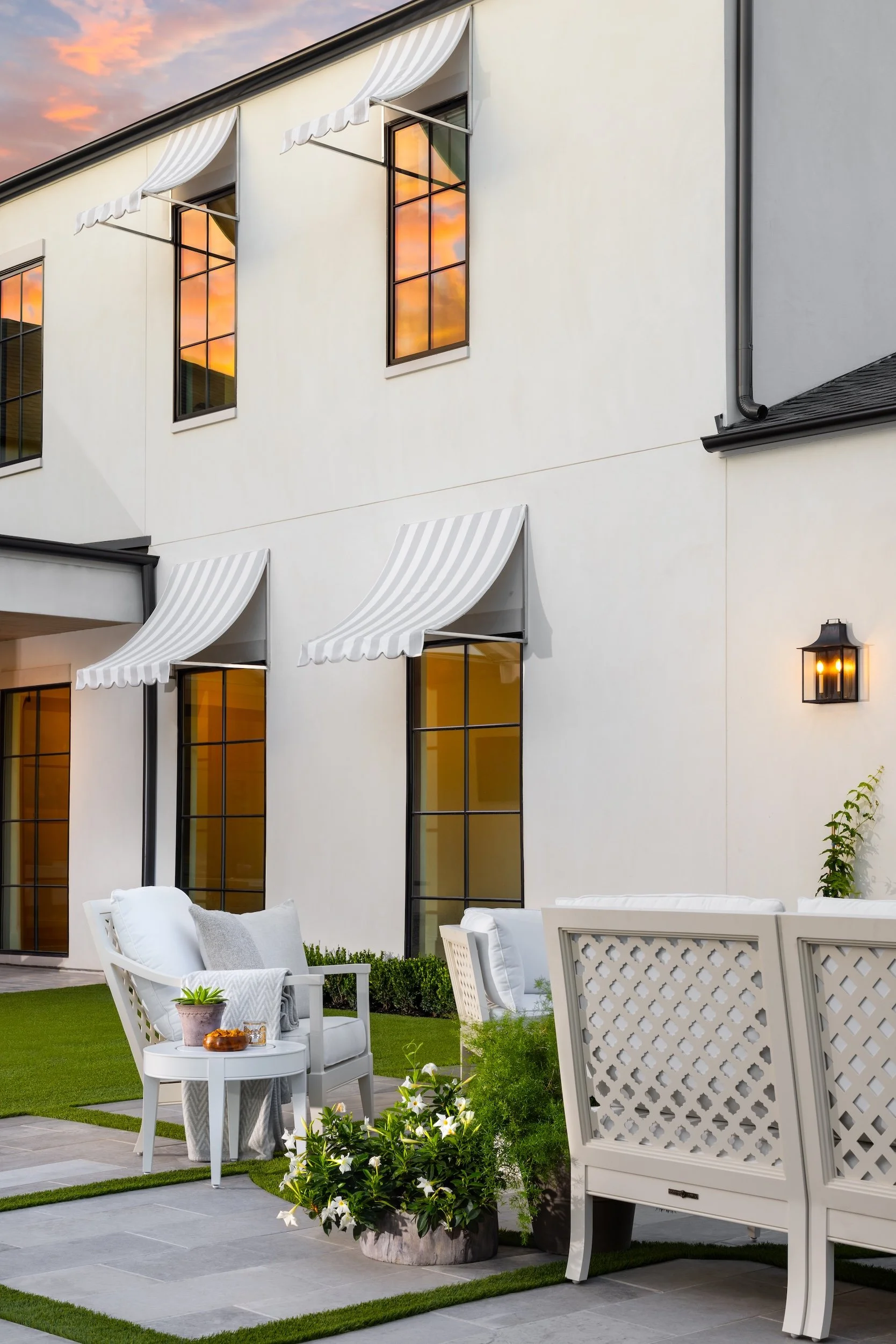 A cozy outdoor patio with white chairs and table, potted plants with white flowers, green grass, and a white house with large windows reflected sunset sky.