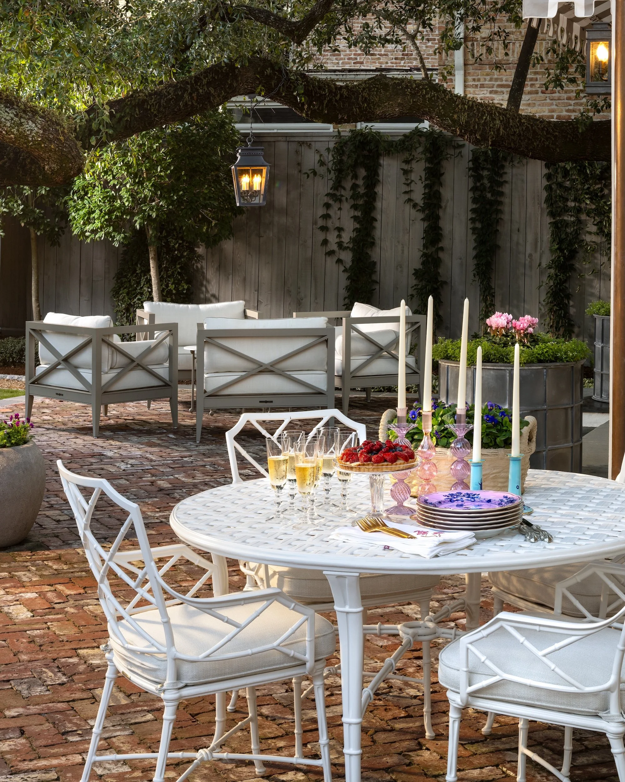 An outdoor patio scene with a round white table set for a meal and white chairs surrounding it. The table has a floral centerpiece with pink flowers, candlesticks, and champagne glasses. In the background, there is a sitting area with white cushioned chairs and sofas, a wooden fence with climbing plants, and a large tree with a hanging lantern. A brick-paved ground and some potted plants are also visible.