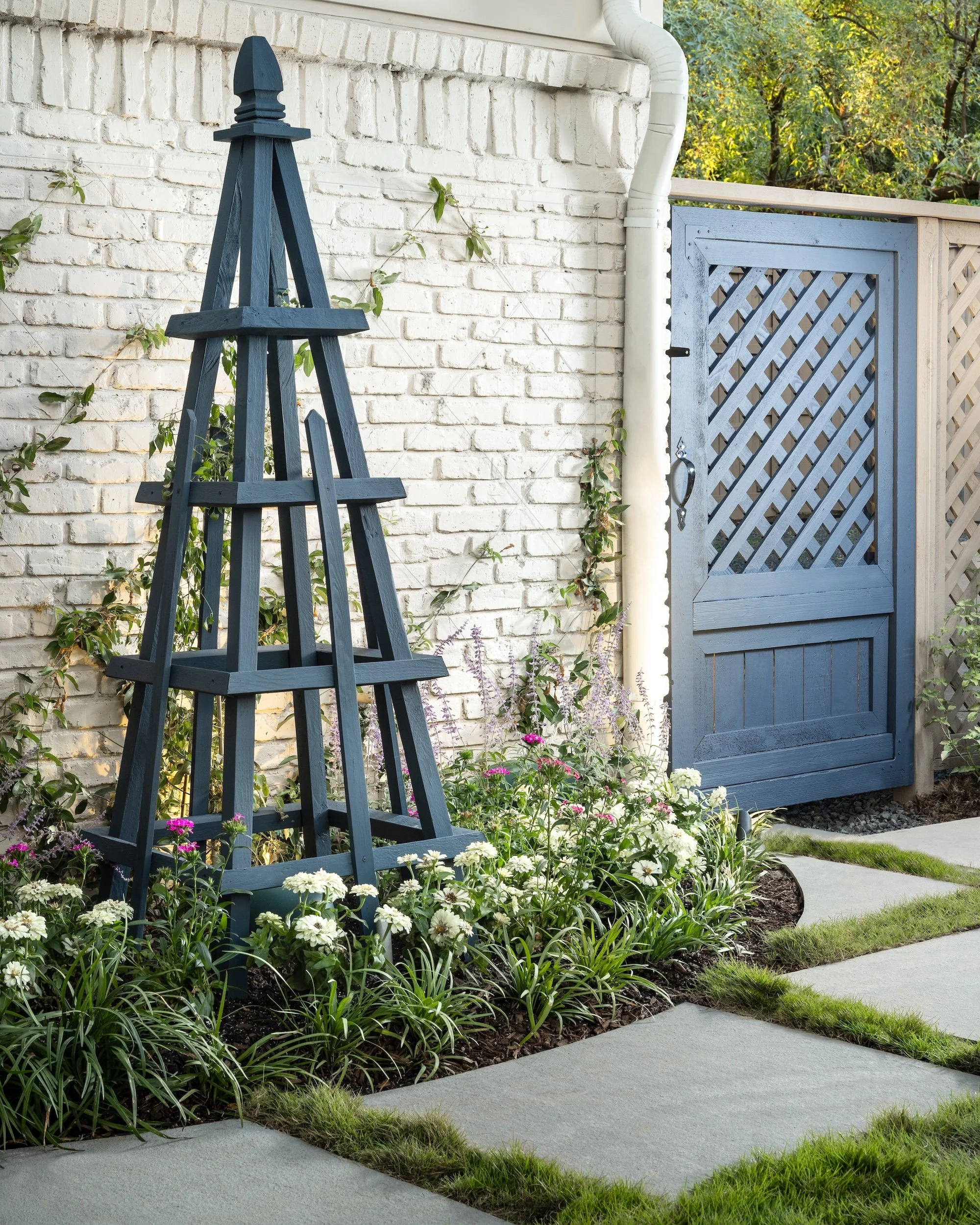 A garden path with concrete stepping stones and green grass, a flower bed with white and purple flowers, a blue garden trellis, a white brick wall with plants climbing, and a blue garden gate with lattice design, surrounded by trees.
