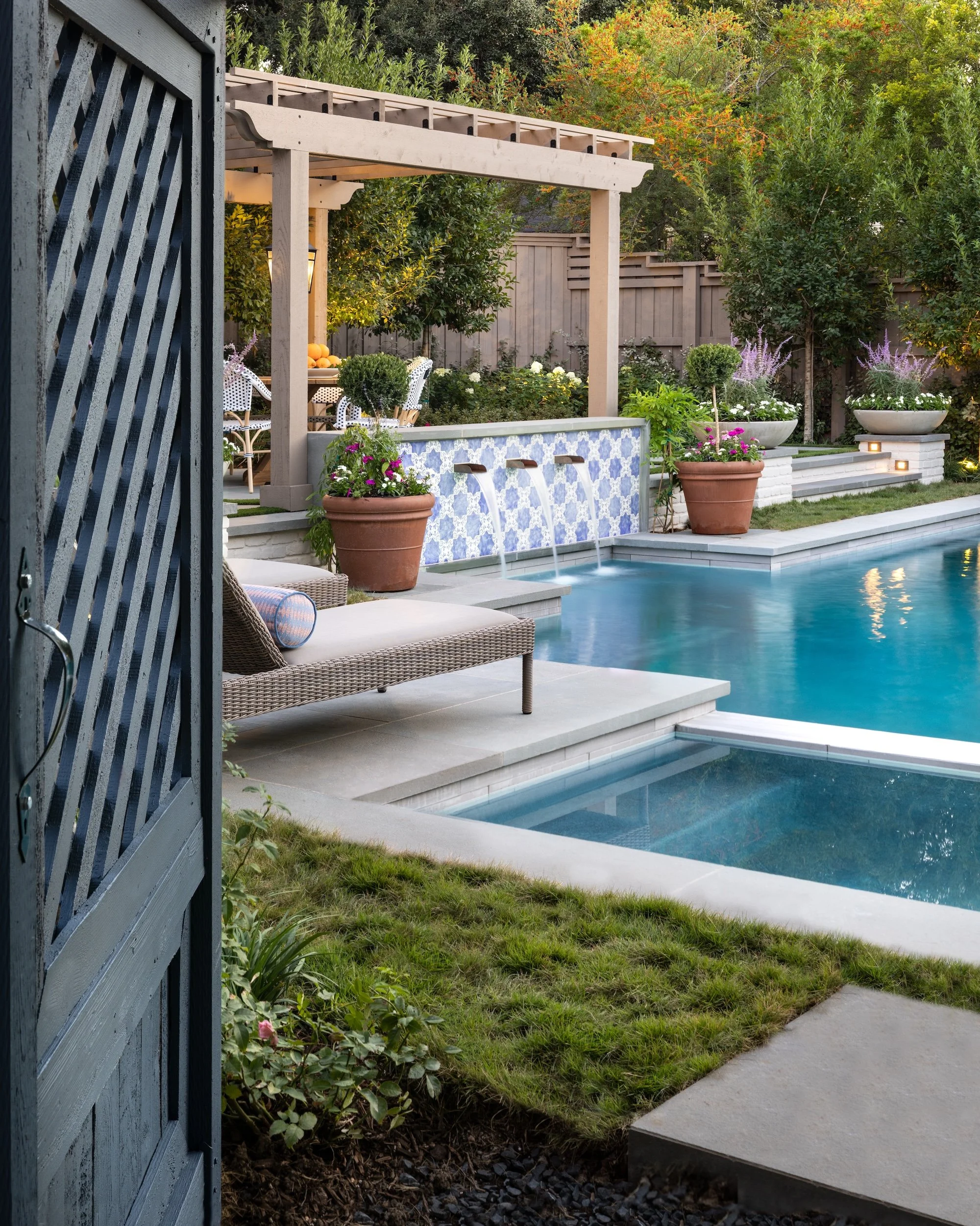 A backyard garden view with a blue-tiled water feature with three spouts, surrounded by potted plants and flowers, a seating area with a table and chairs, and a wooden pergola, all enclosed by a wooden fence and greenery.