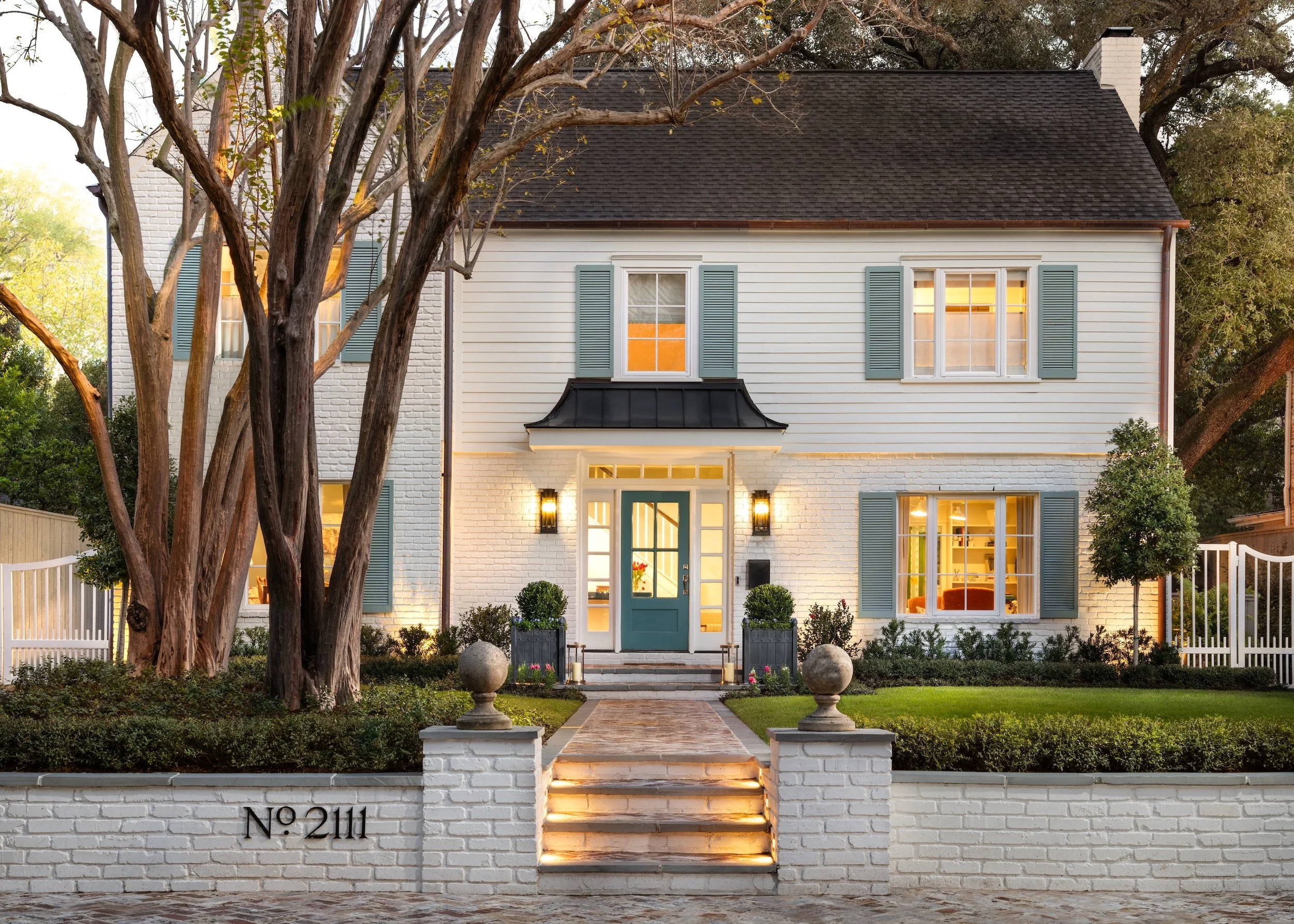 Front view of a two-story white house with teal window shutters, a black metal awning over the front door, and a white brick fence with steps leading to the entrance. There are trees in front of the house and exterior lights illuminating the entrance.