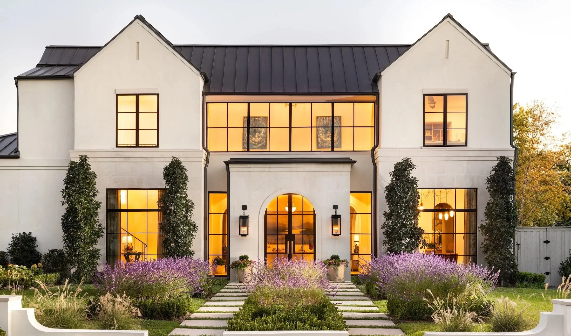 Front view of a modern house with large glass windows, a dark metal roof, and well-maintained garden with pathways, trees, and purple flowers.
