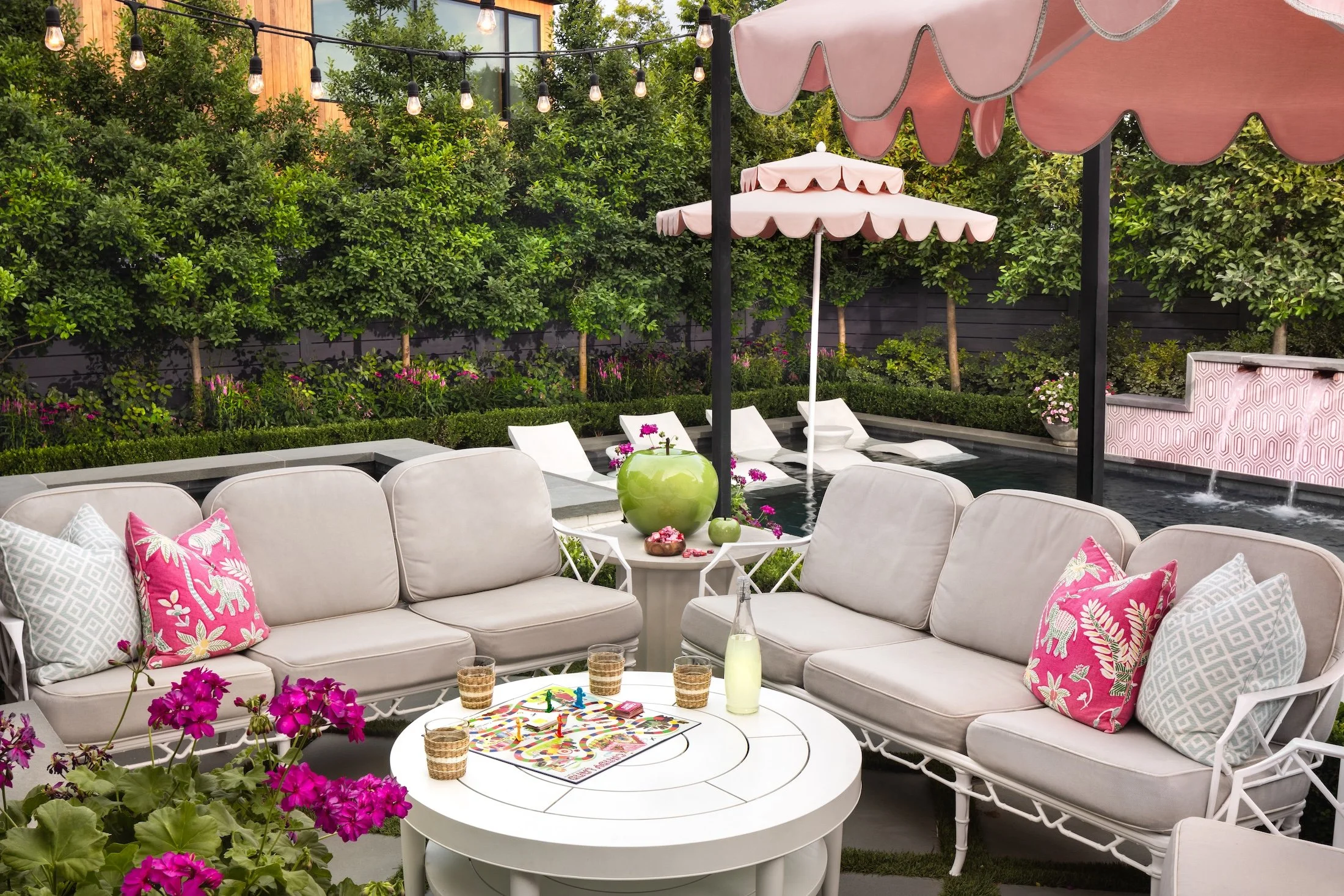 A lively outdoor backyard party scene with white patio furniture, pink and white umbrellas, and pink cushions. There are string lights overhead, surrounded by lush greenery and flowers. On the table, there is a game, drinks, and a decorative green vase with flowers.