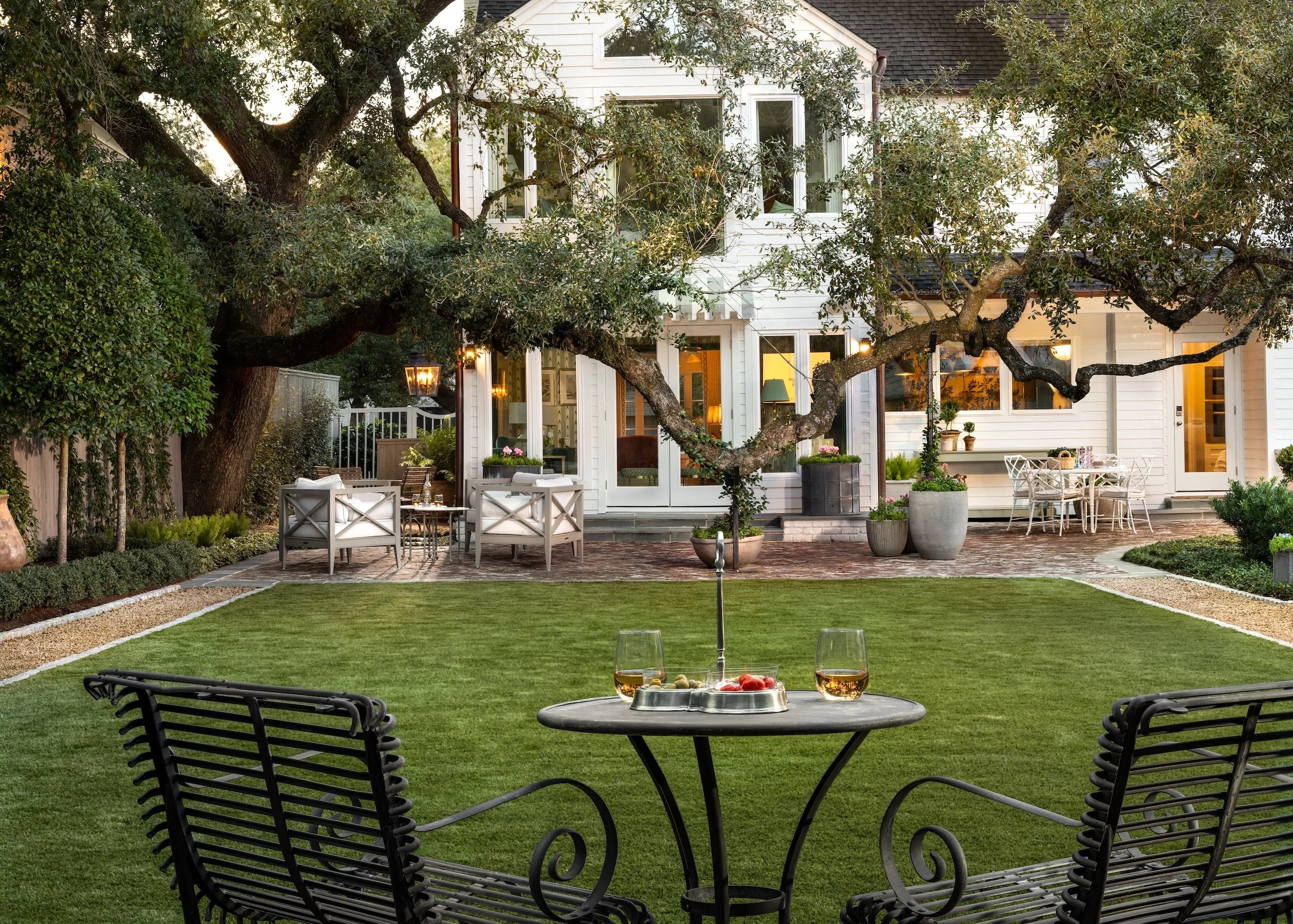 A backyard patio with a table set for drinks, overlooking a yard with a house in the background, surrounded by trees and landscaping.