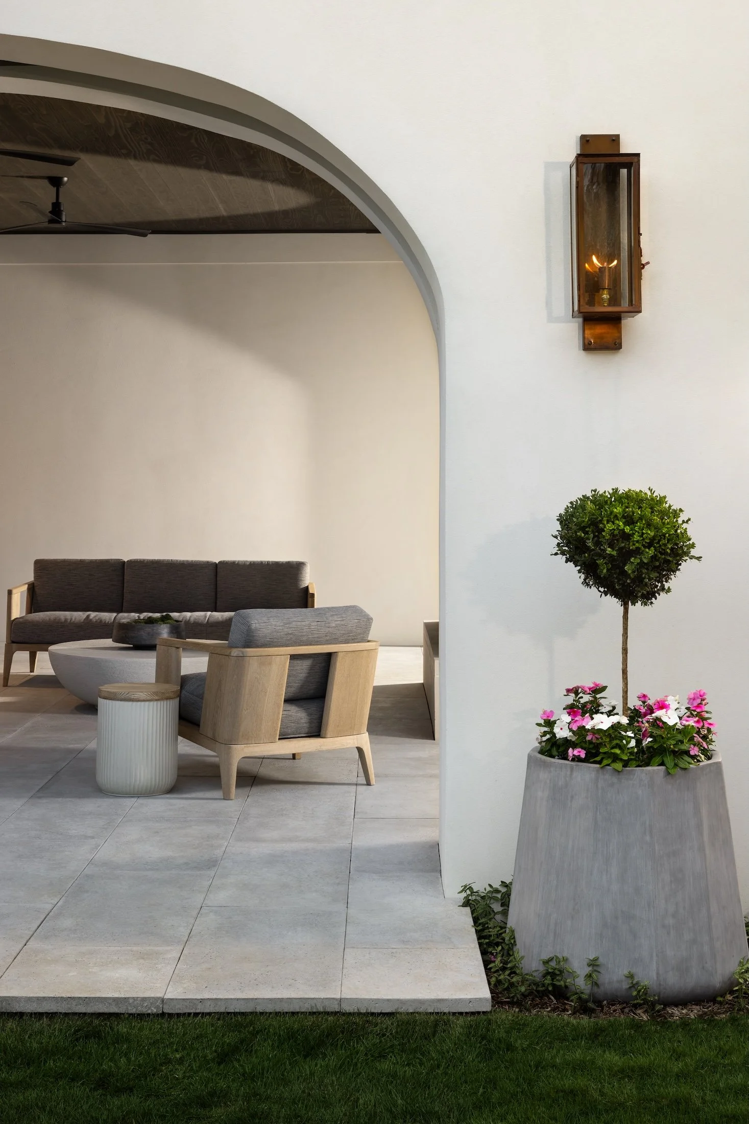 Outdoor patio area with modern furniture, a large potted plant with pink flowers, and a wall-mounted light fixture with a flickering candle inside.