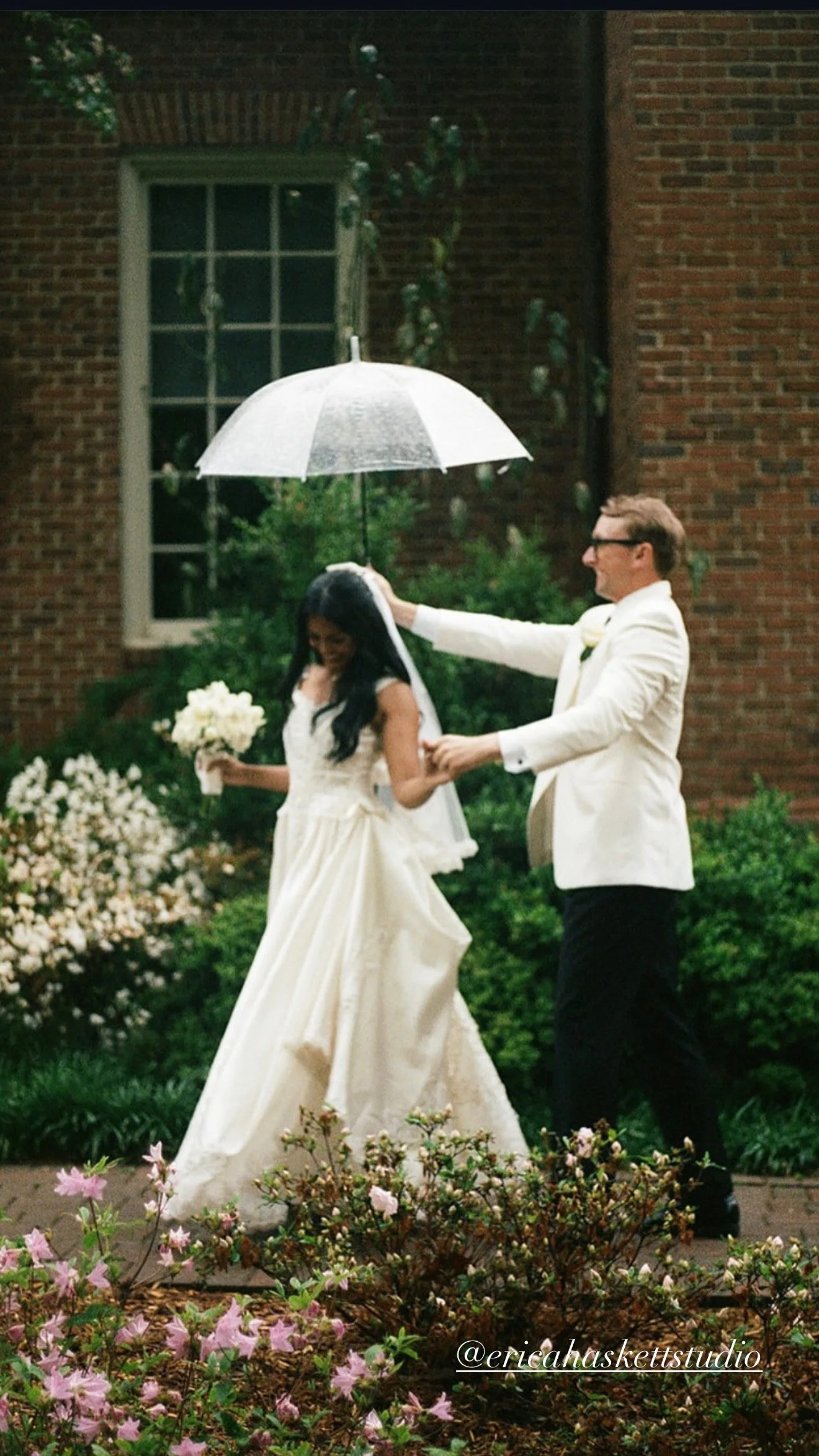 A bride and groom walk outdoors in a garden during a rain shower. The groom holds an umbrella over the bride, who is holding a bouquet of white flowers. The bride is wearing a white wedding gown, and the groom is dressed in a white tuxedo jacket. The background includes a brick building, green shrubbery, and blooming pink and white flowers.
