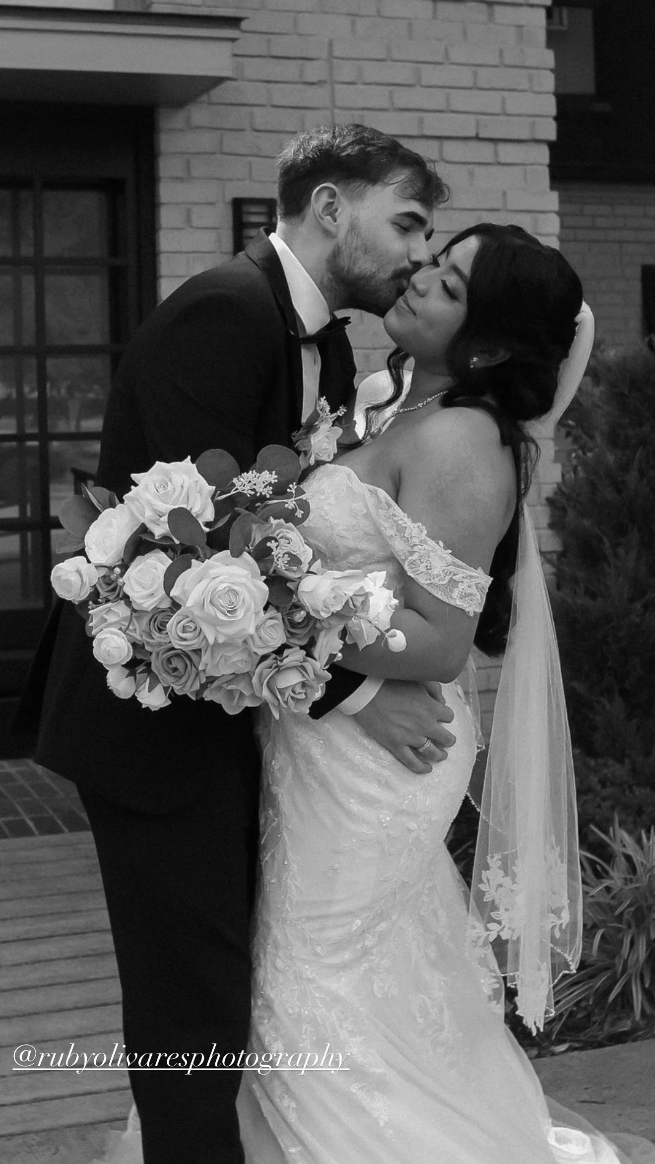 A black and white photograph of a bride and groom on their wedding day. The groom is kissing the bride on the cheek while she smiles with closed eyes. The groom wears a tuxedo, and the bride wears a lace wedding dress and holds a large bouquet of roses and greenery. They are standing outdoors in front of a brick building.
