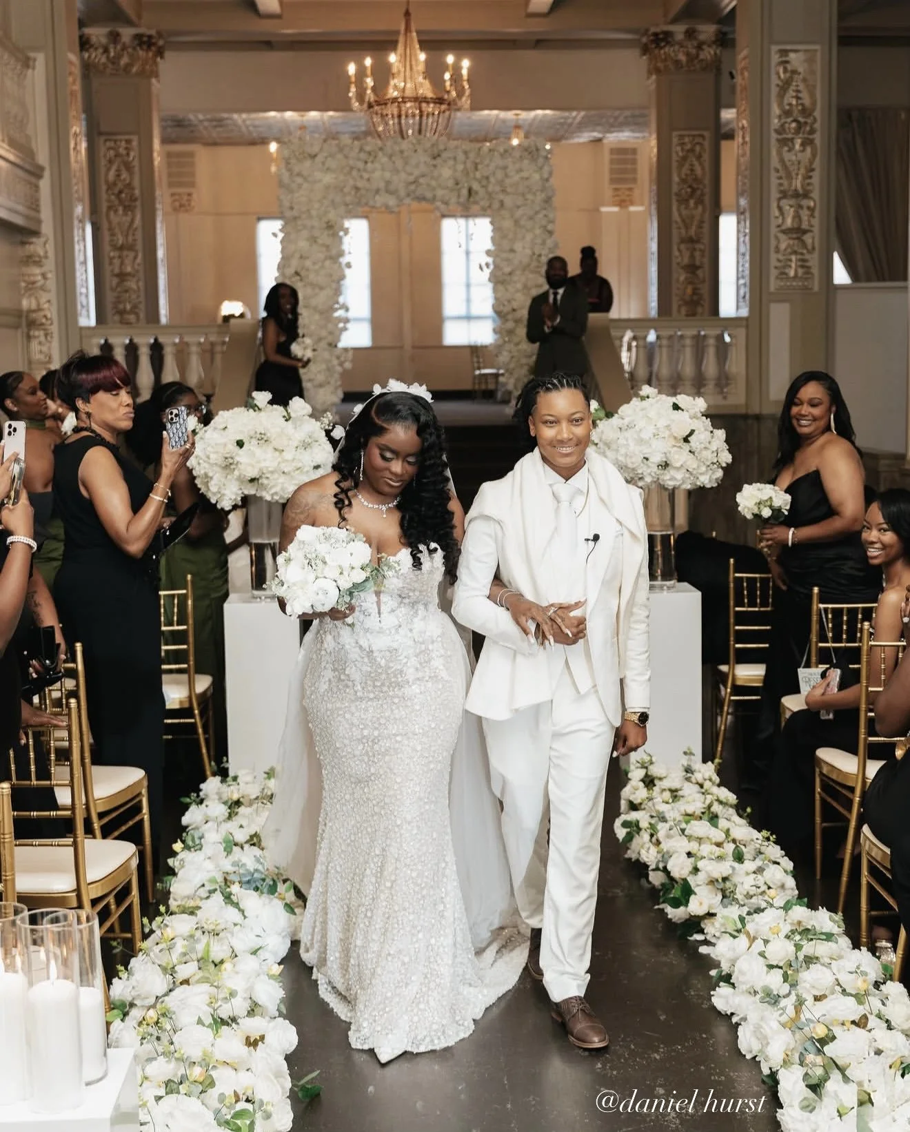 A wedding ceremony with two brides walking down the aisle decorated with white flowers. The bride on the left is wearing a white lace wedding gown, holding a bouquet, and has long, dark, curly hair. The bride on the right is dressed in a white suit with a matching shirt, brown shoes, and her hair styled in braids. Guests in black dresses are seated and taking photos, with some standing and smiling. The backdrop features a floral arch and a grand chandelier in a luxurious hall with ornate decor.
