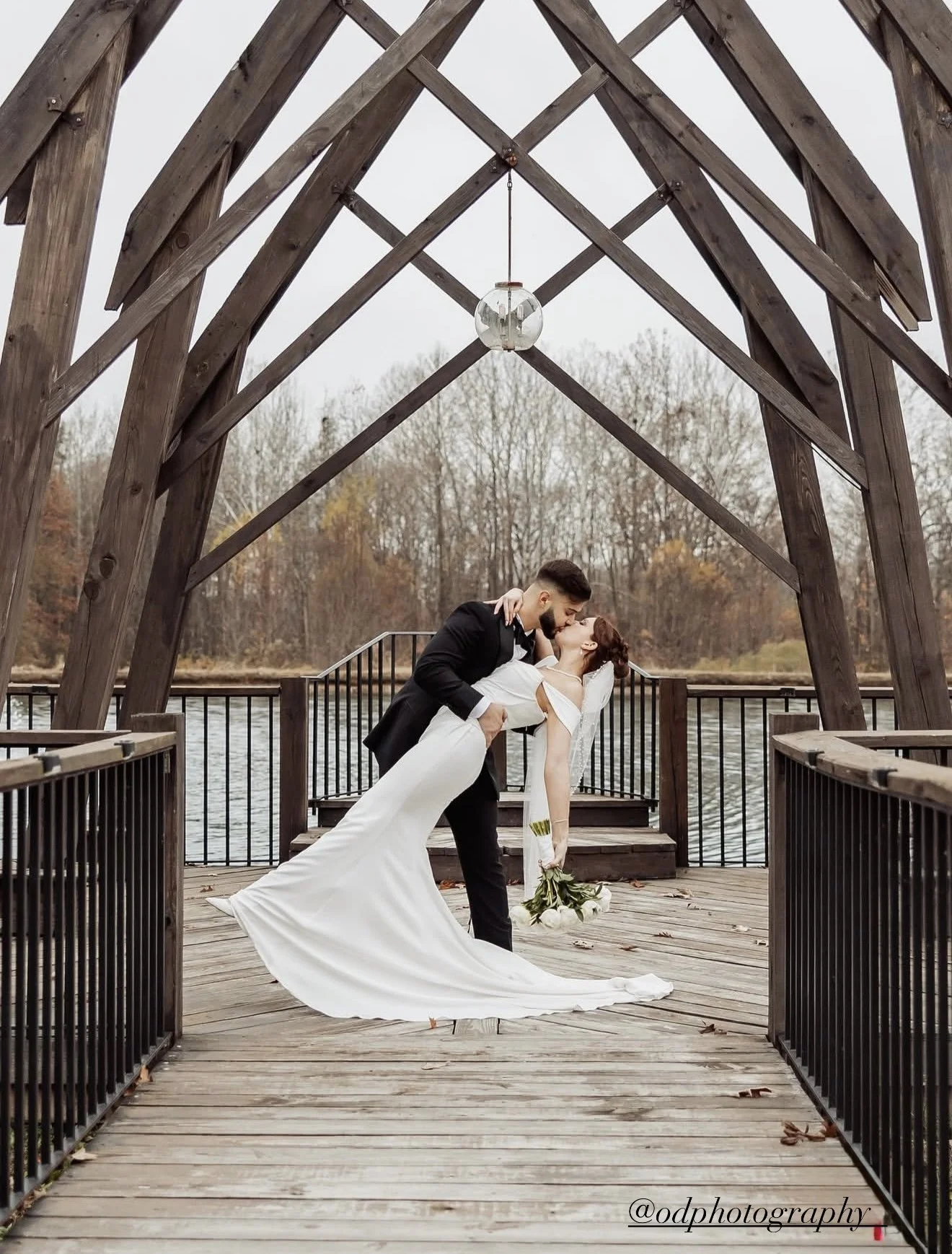 A bride and groom sharing a kiss on a wooden dock with a lake and trees in the background, under a wooden arch with a hanging light fixture.