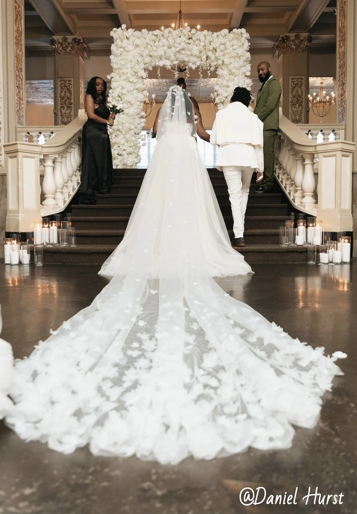 A wedding ceremony with two brides walking down the aisle decorated with white flowers. The bride on the left is wearing a white lace wedding gown, holding a bouquet, and has long, dark, curly hair. The bride on the right is dressed in a white suit with a matching shirt, brown shoes, and her hair styled in braids. Guests in black dresses are seated and taking photos, with some standing and smiling. The backdrop features a floral arch and a grand chandelier in a luxurious hall with ornate decor.