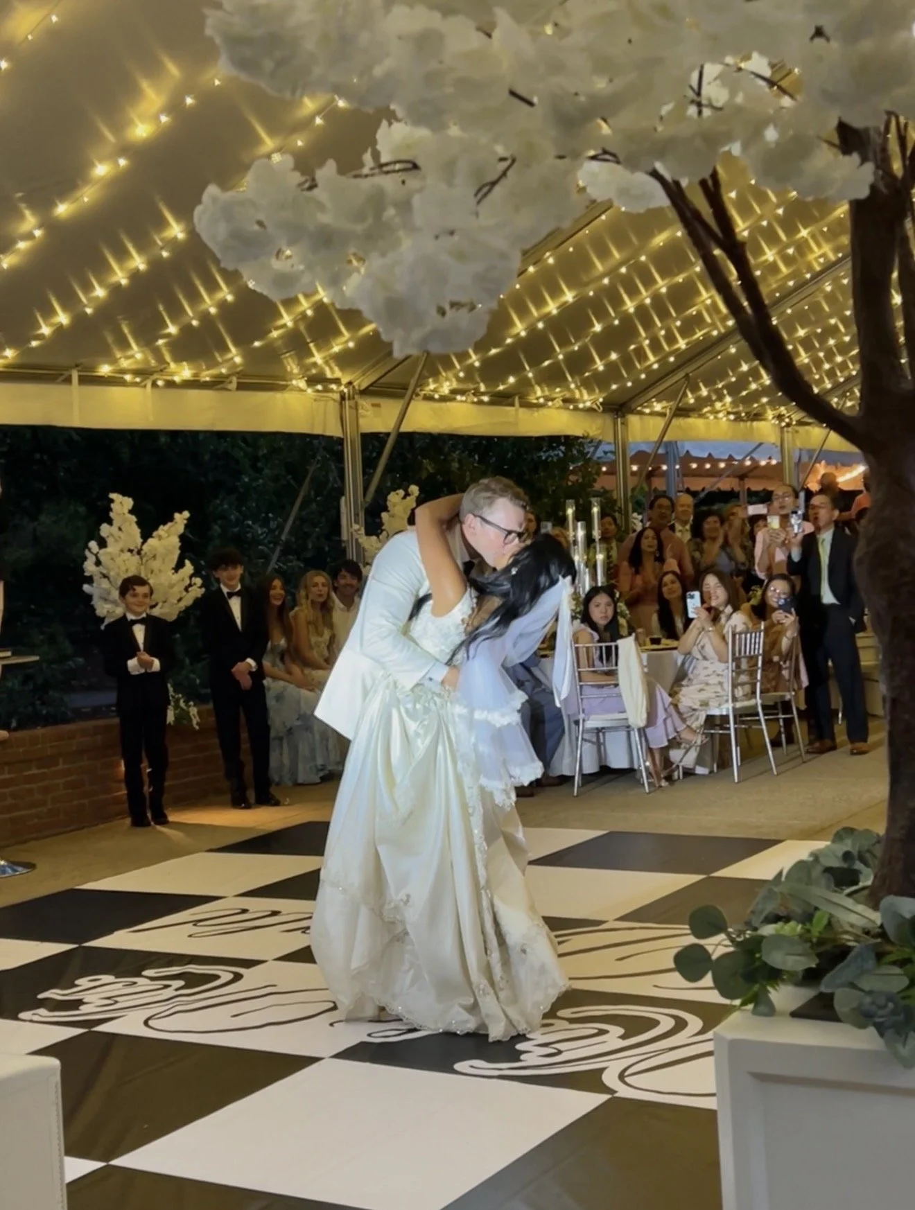 A bride and groom walk outdoors in a garden during a rain shower. The groom holds an umbrella over the bride, who is holding a bouquet of white flowers. The bride is wearing a white wedding gown, and the groom is dressed in a white tuxedo jacket. The background includes a brick building, green shrubbery, and blooming pink and white flowers.