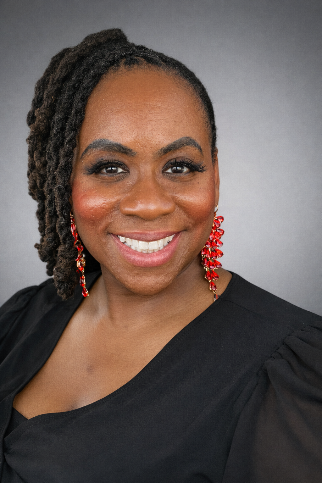 Close-up portrait of a smiling woman with dark curly dreadlocks, wearing red dangling earrings and a black top, against a gray background.