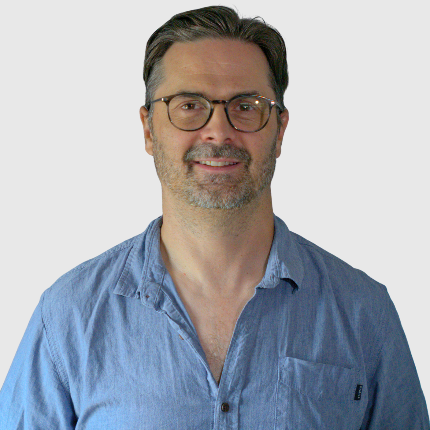 A middle-aged man with gray hair, wearing glasses and a light blue button-up shirt, smiling against a plain white background.