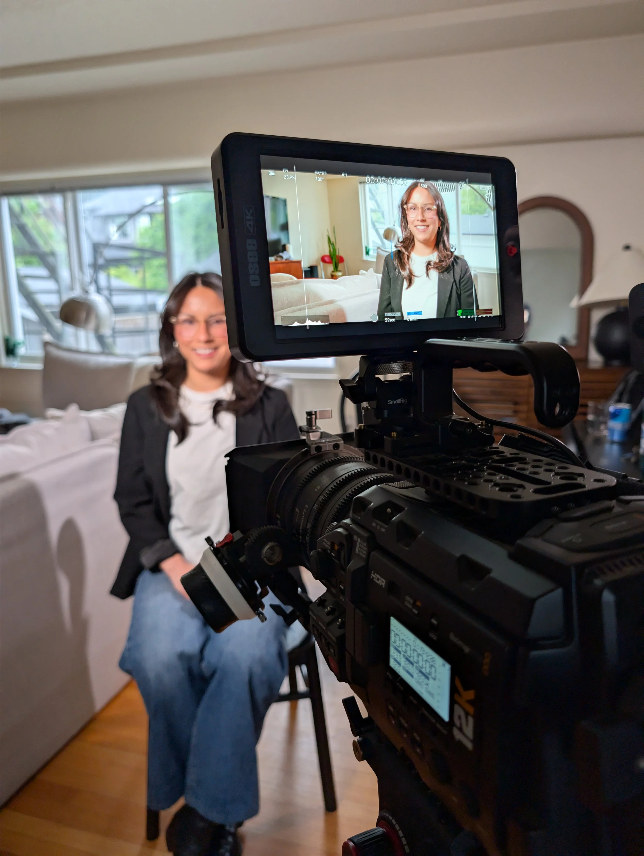 A woman sitting on a chair in a living room, being filmed or photographed with a professional camera on a tripod. She is smiling, wearing glasses, a black blazer, and blue jeans. The background shows a window with greenery outside, and some furniture including a lamp and a dresser.