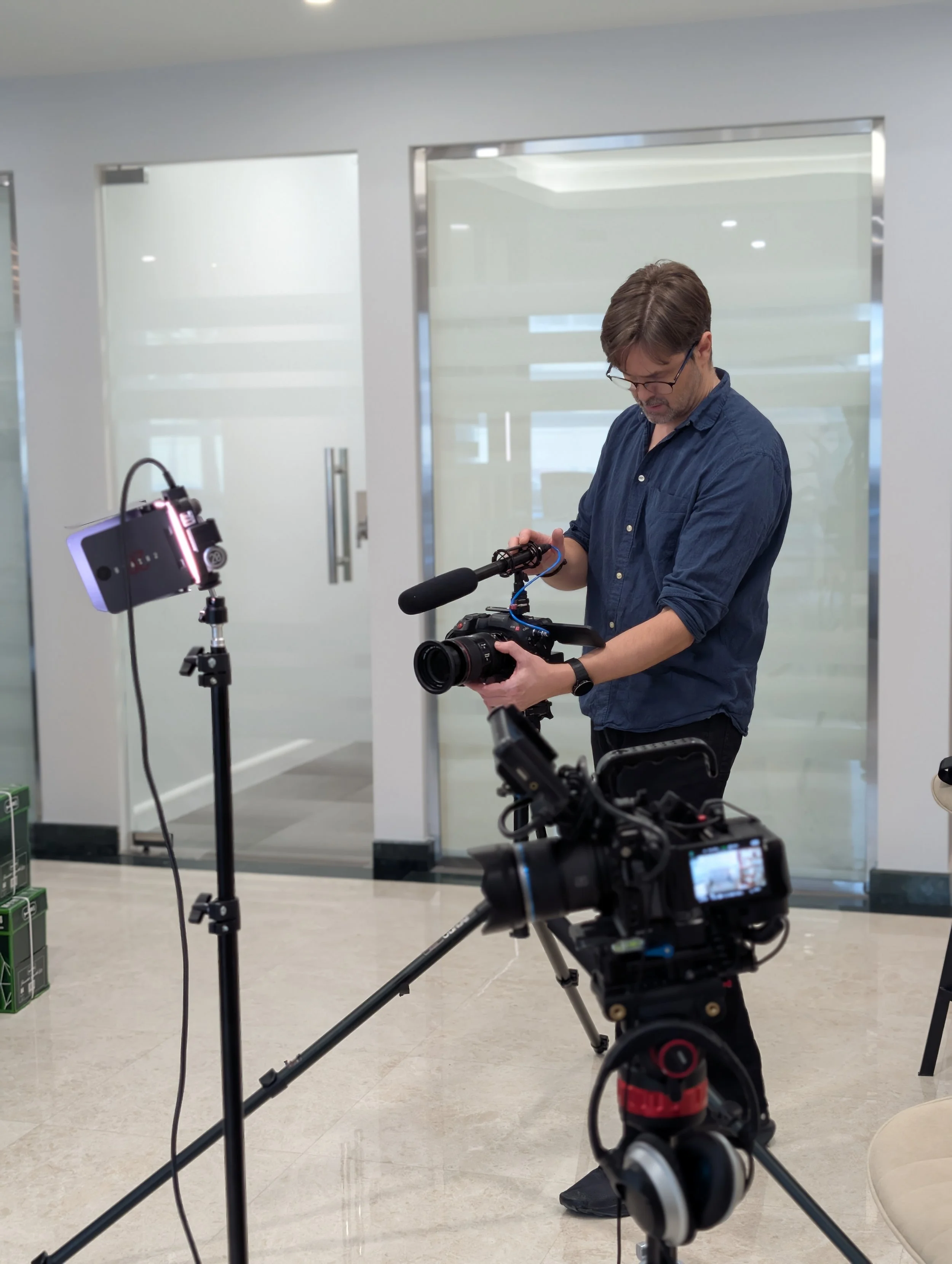A man wearing glasses and a blue shirt is holding a professional video camera. He is in an indoor setting with white walls and glass doors, surrounded by filming equipment, including a microphone and additional cameras.