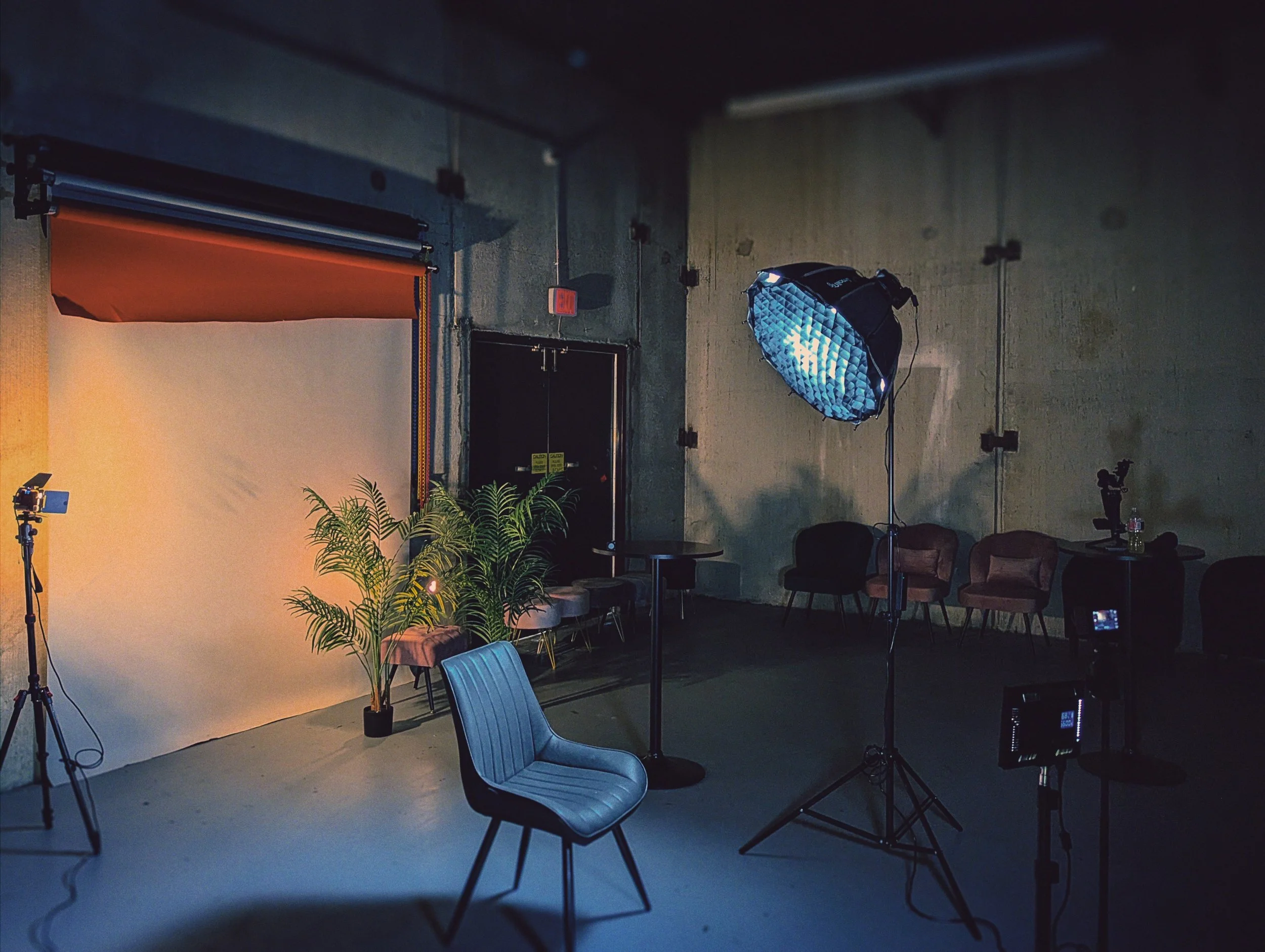An indoor photography studio setup with a black backdrop, a softbox light on a stand, chairs arranged in front of the backdrop, and a camera on a tripod pointing towards the backdrop. There are plants and other equipment visible in the space.