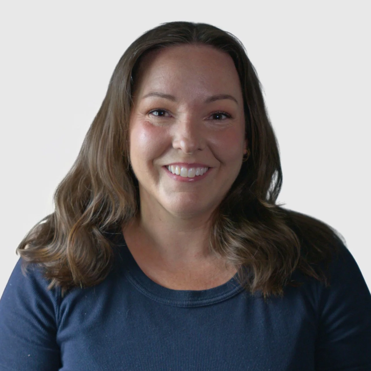 A woman with long wavy brown hair smiling, wearing a navy blue top with a grey background.