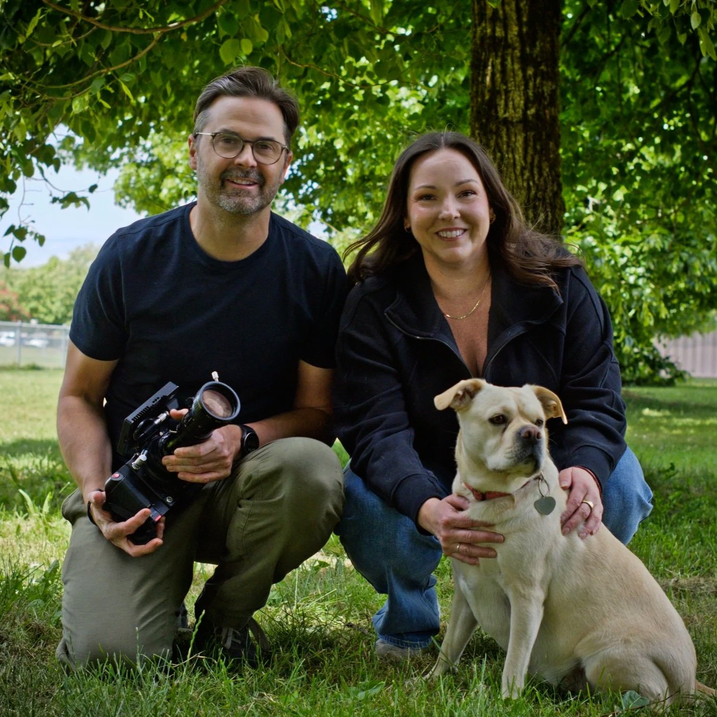 A man with glasses and a beard holding a camera kneels next to a woman with long brown hair, who is holding a tan bulldog. They are outdoors in a green park with trees in the background, both smiling.