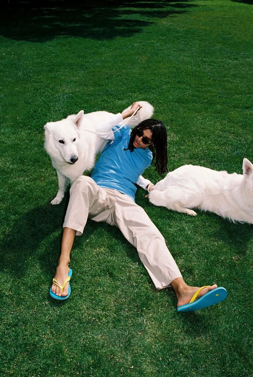Man with dreadlocks and sunglasses lying on green grass, holding two white dogs, one sitting and one lying down, wearing casual clothing and flip-flops.