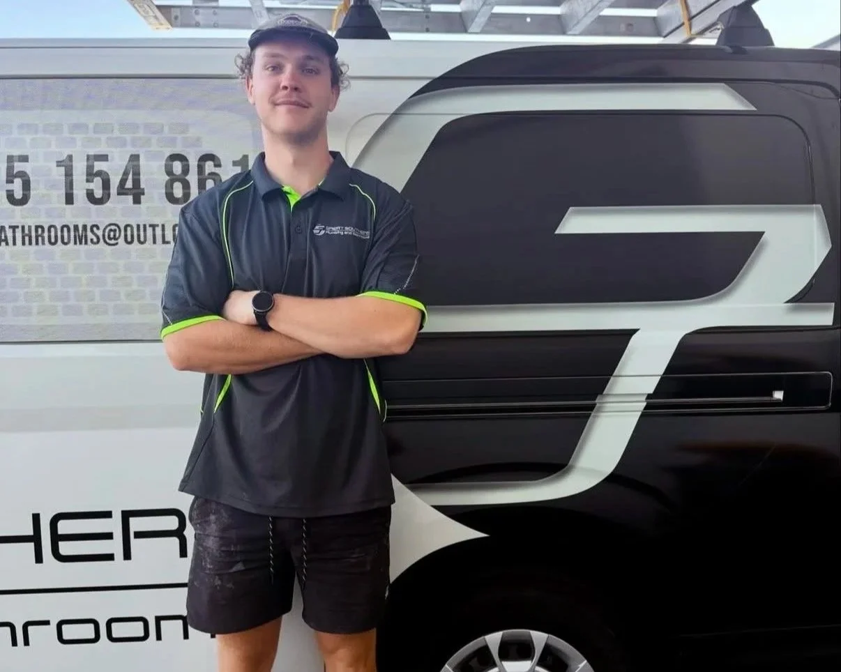 Young man in black and neon green polo shirt standing with arms crossed in front of a black and white company van with logo and contact information.