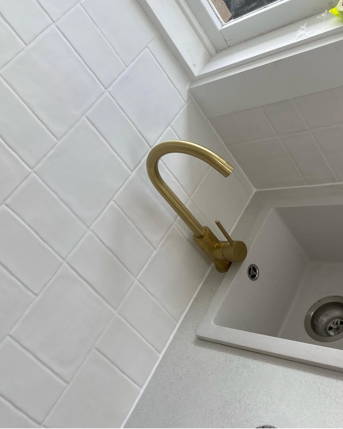 Close-up of a gold kitchen faucet above a white sink, with white tiled backsplash and a window in the background.