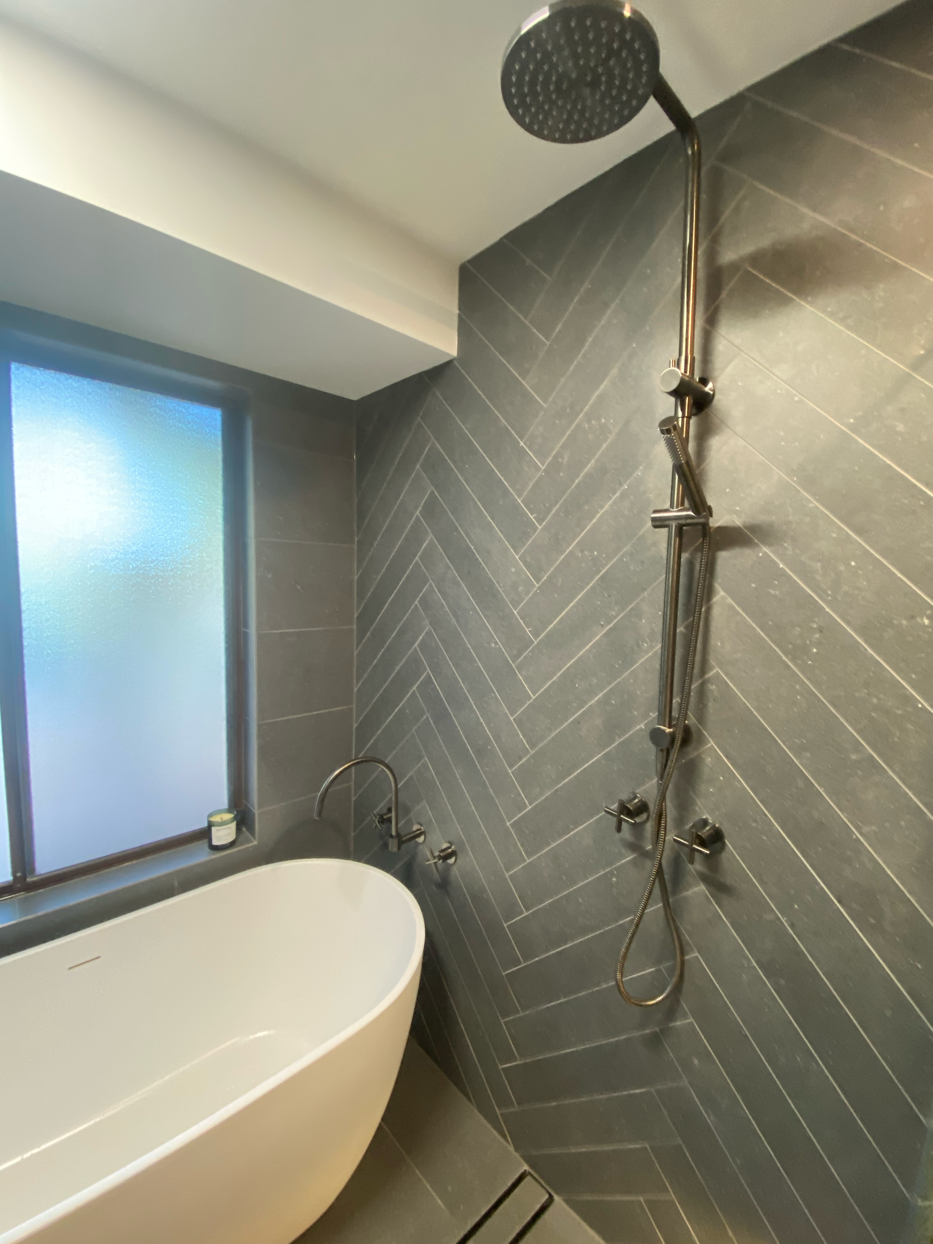 A modern shower area with black tiled walls, a ceiling-mounted showerhead, a handheld showerhead, and several controls. There is a window with frosted glass in the background.