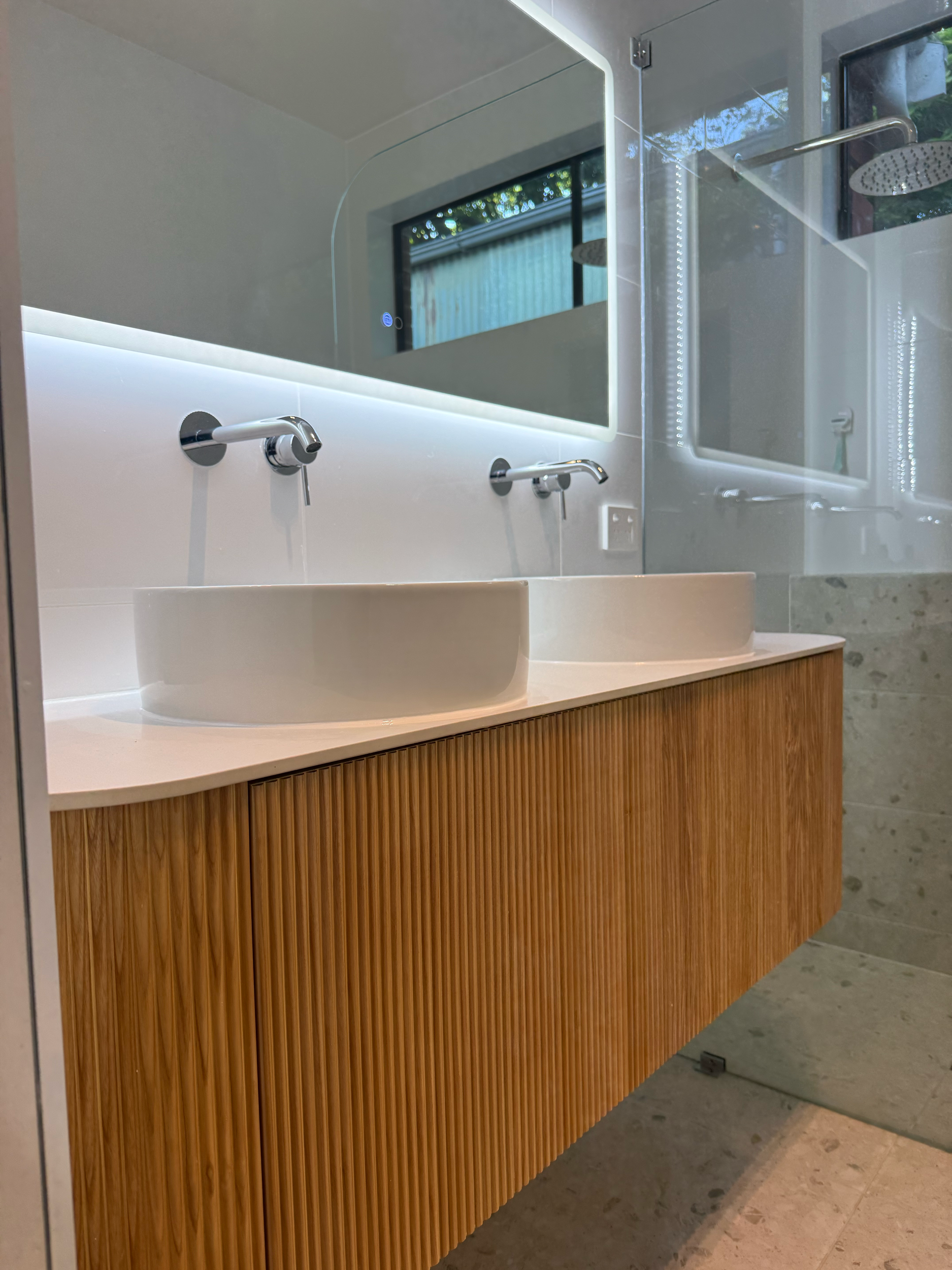 The image shows a modern, minimalist bathroom with a white vanity that includes two white sinks and chrome faucets. Above the vanity is a large mirror, and a wooden slatted cabinet is positioned to the right side. The floor consists of gray stone til