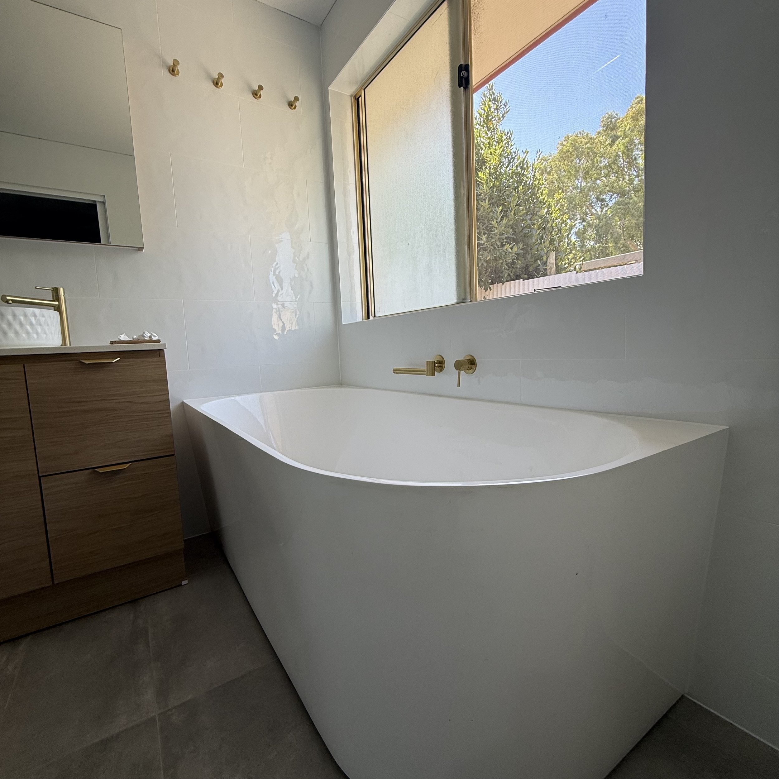 Modern bathroom with large window overlooking trees, white bathtub, wooden vanity, gold faucet, and minimal decor.