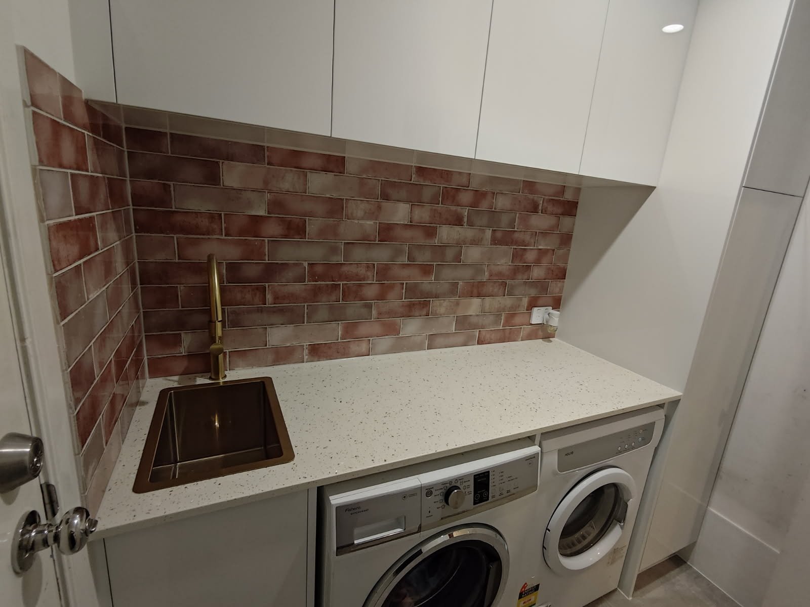 A small laundry area with a speckled white countertop, brick wall backsplash, a mini stainless steel sink with a gold faucet, white upper cabinets, a washing machine, and a dryer beneath the countertop.
