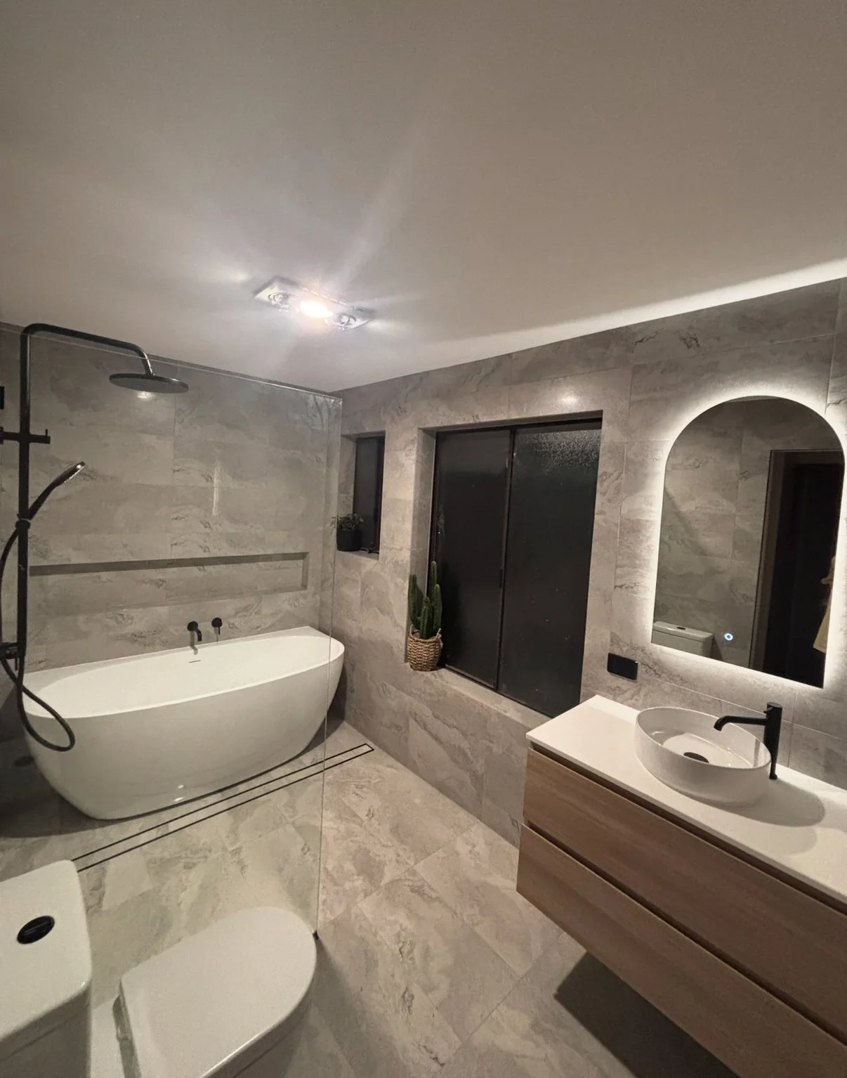 Modern bathroom with a freestanding bathtub, a walk-in shower, a vanity with a vessel sink, a mirror with backlit lighting, and decorative plants.