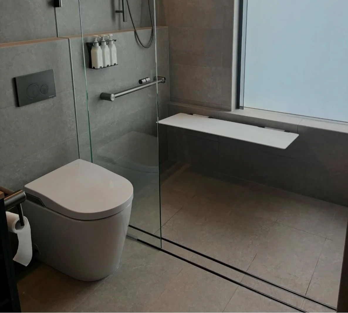Modern bathroom with gray tile walls and floor, a white toilet, a glass shower enclosure with a rain showerhead, a fold-down bench, and a window.